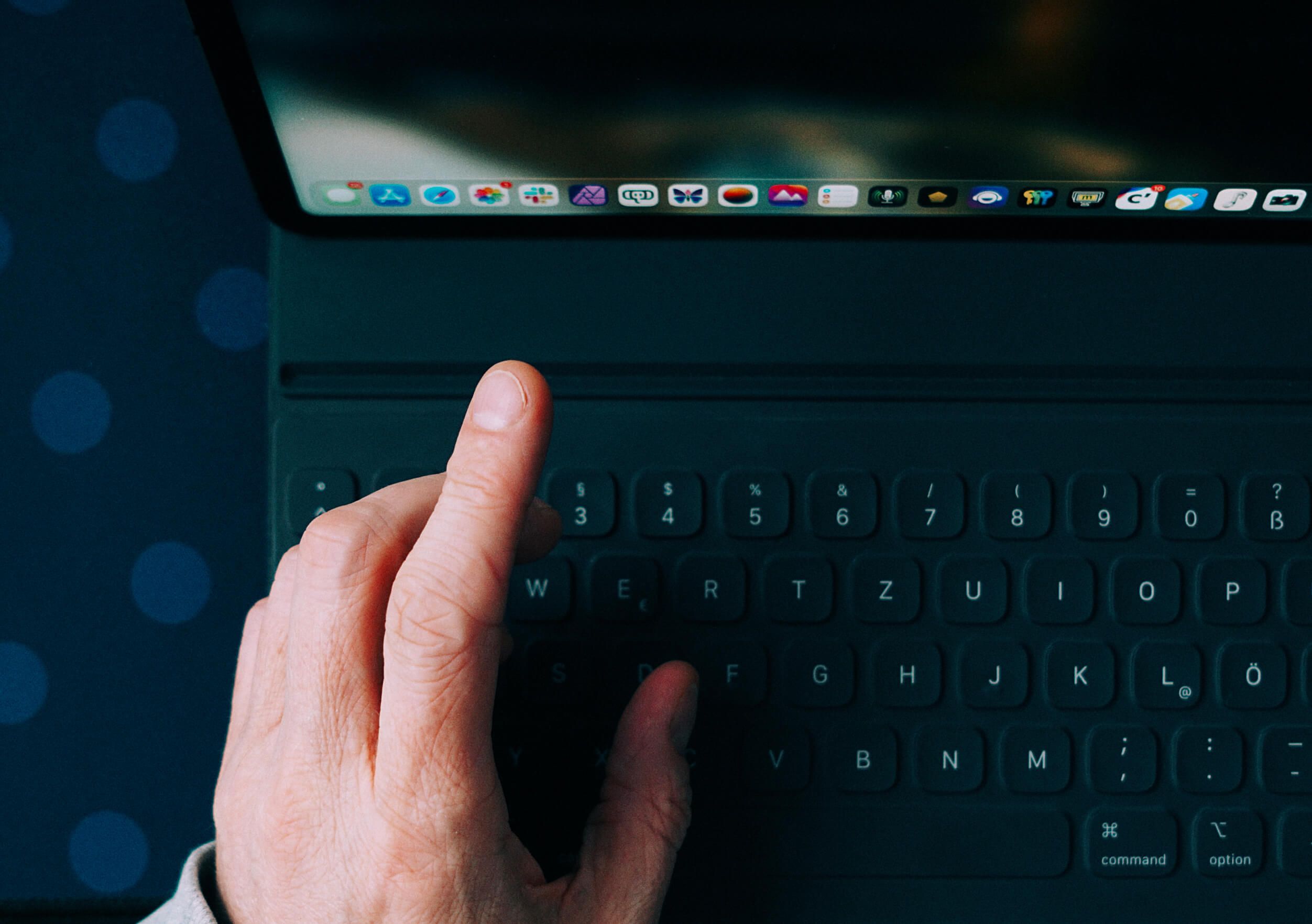 A person's left hand hovers above the dark keyboard of a tablet or laptop with the screen partially visible, showing several app icons along the top edge.