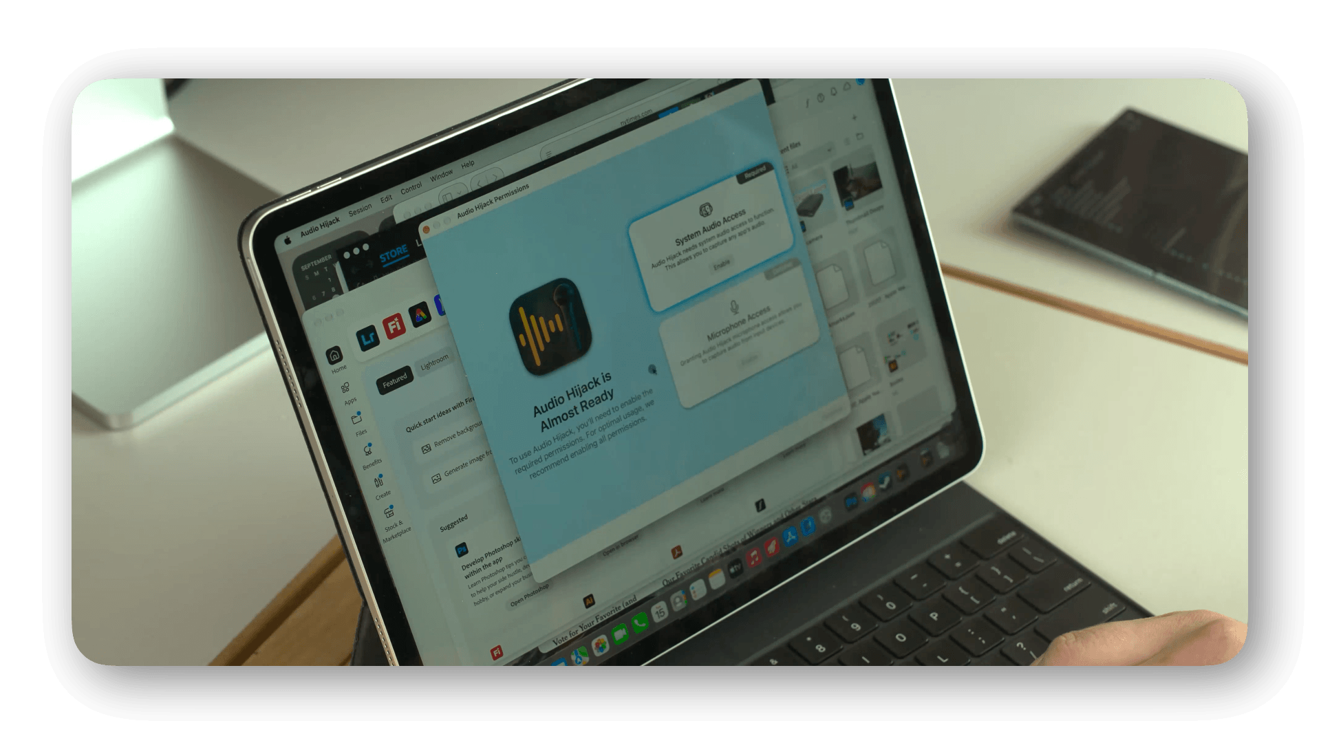 A close-up of a laptop screen showing a permission popup for 'Audio Hijack' software, asking for system audio and microphone access. The laptop is placed on a desk with a hand visible on the keyboard, and a tablet is seen in the background.