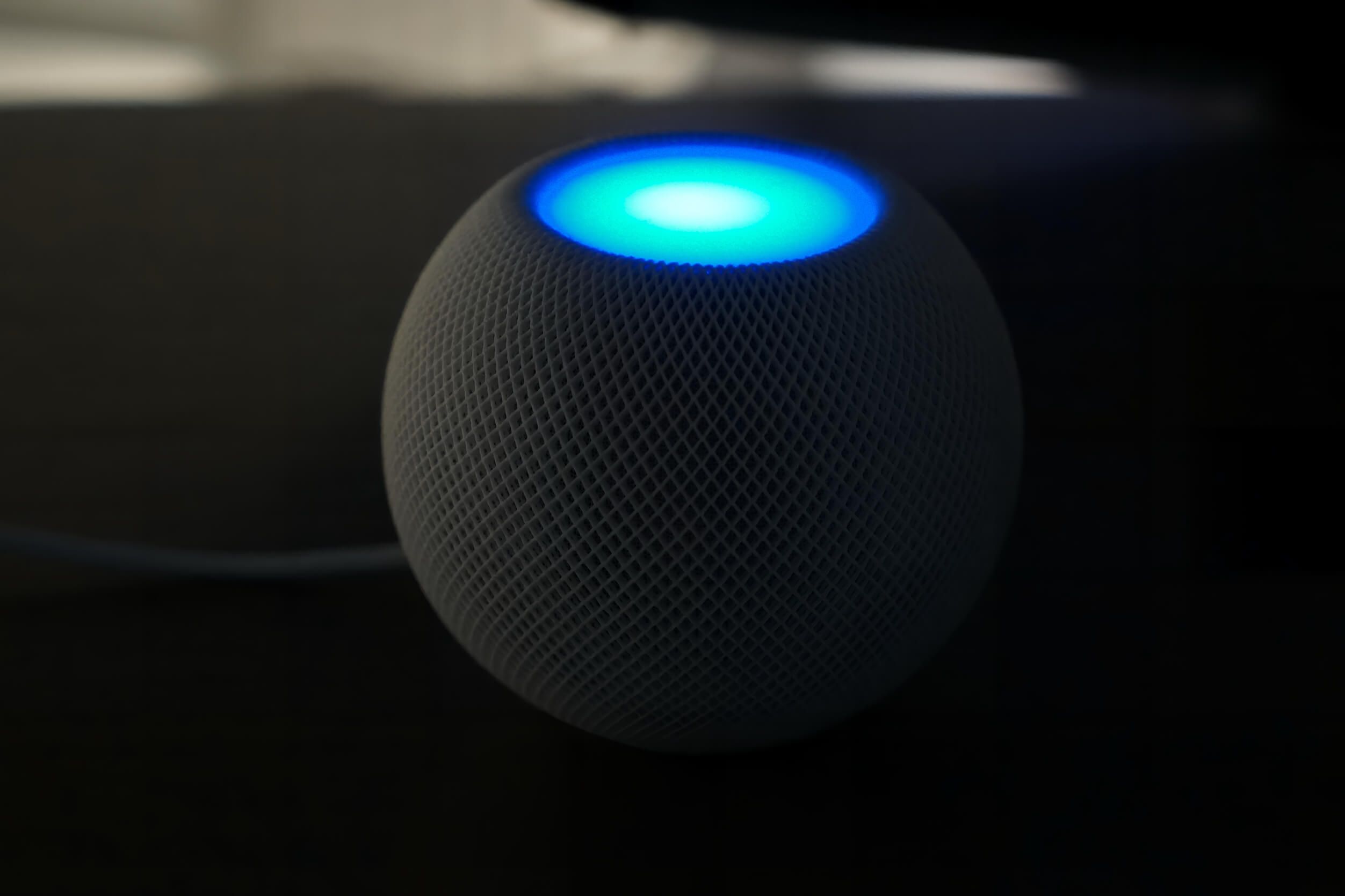 A spherical smart speaker with a mesh exterior and a glowing blue light on top sits on a dark surface, connected by a power cord.