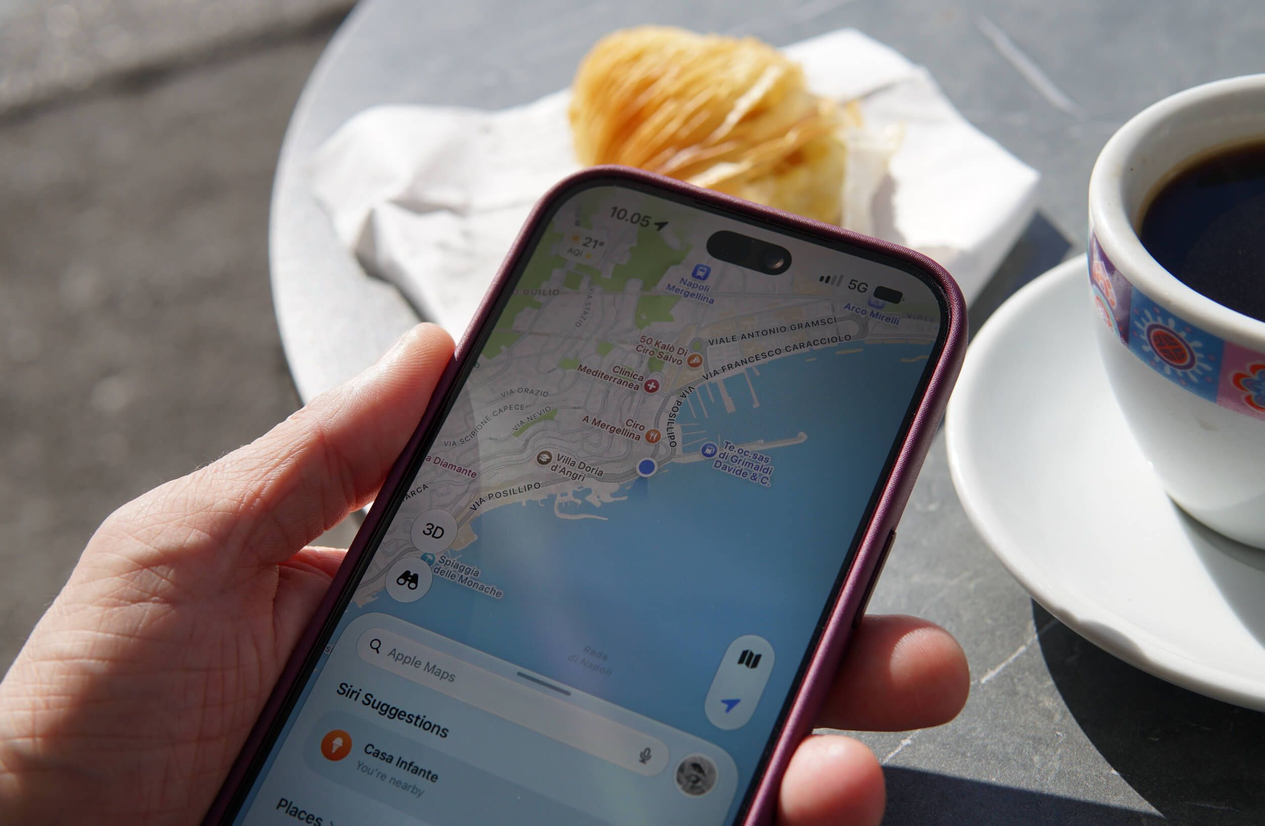 A hand holding a smartphone displaying a map application of a coastal area, with a plate of pastry and a cup of black coffee on a gray table surface nearby.