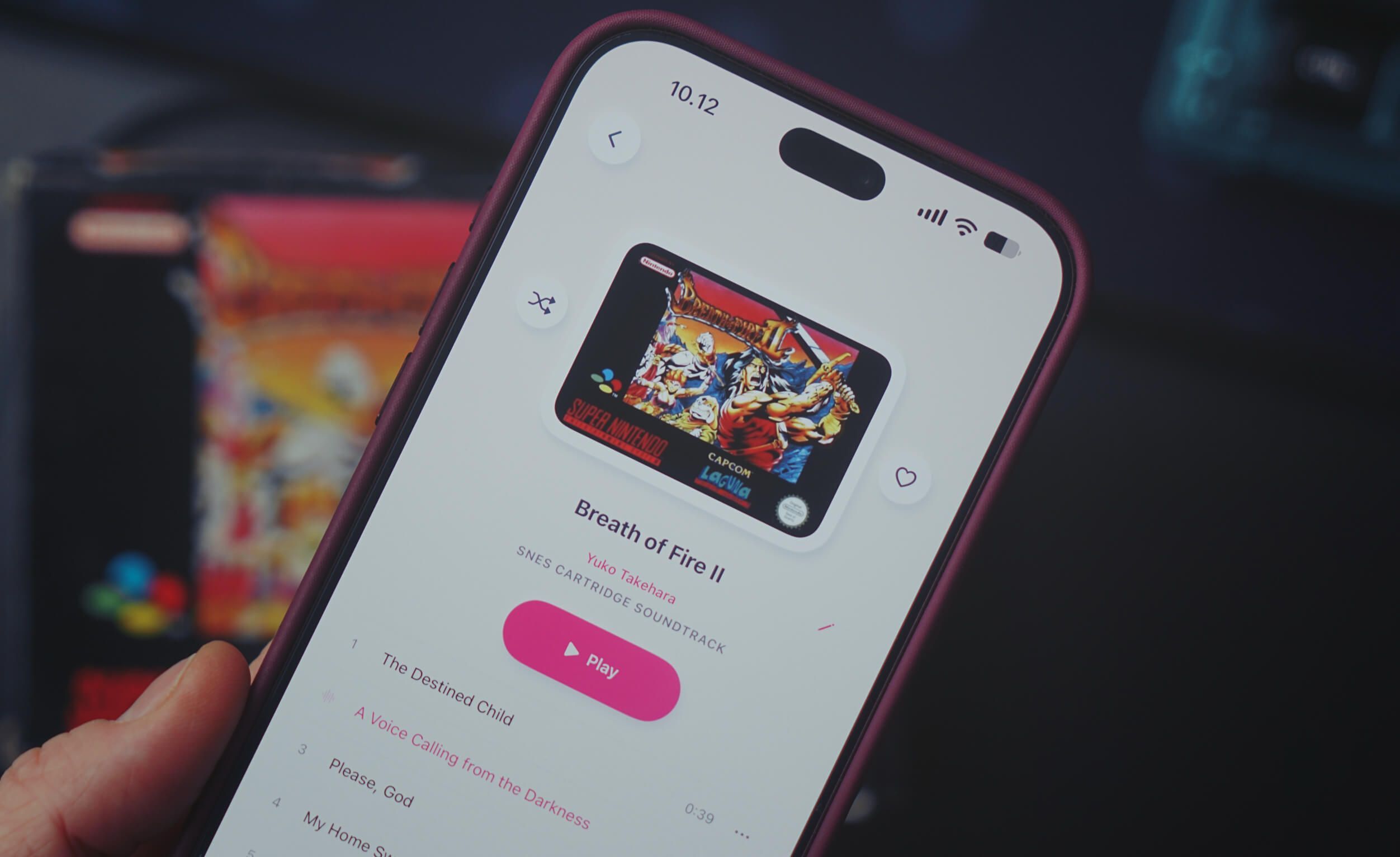A close-up of a hand holding a smartphone with a music app open, displaying the soundtrack for the Super Nintendo game "Breath of Fire II". The screen shows the game cover art, track list, and a pink play button.