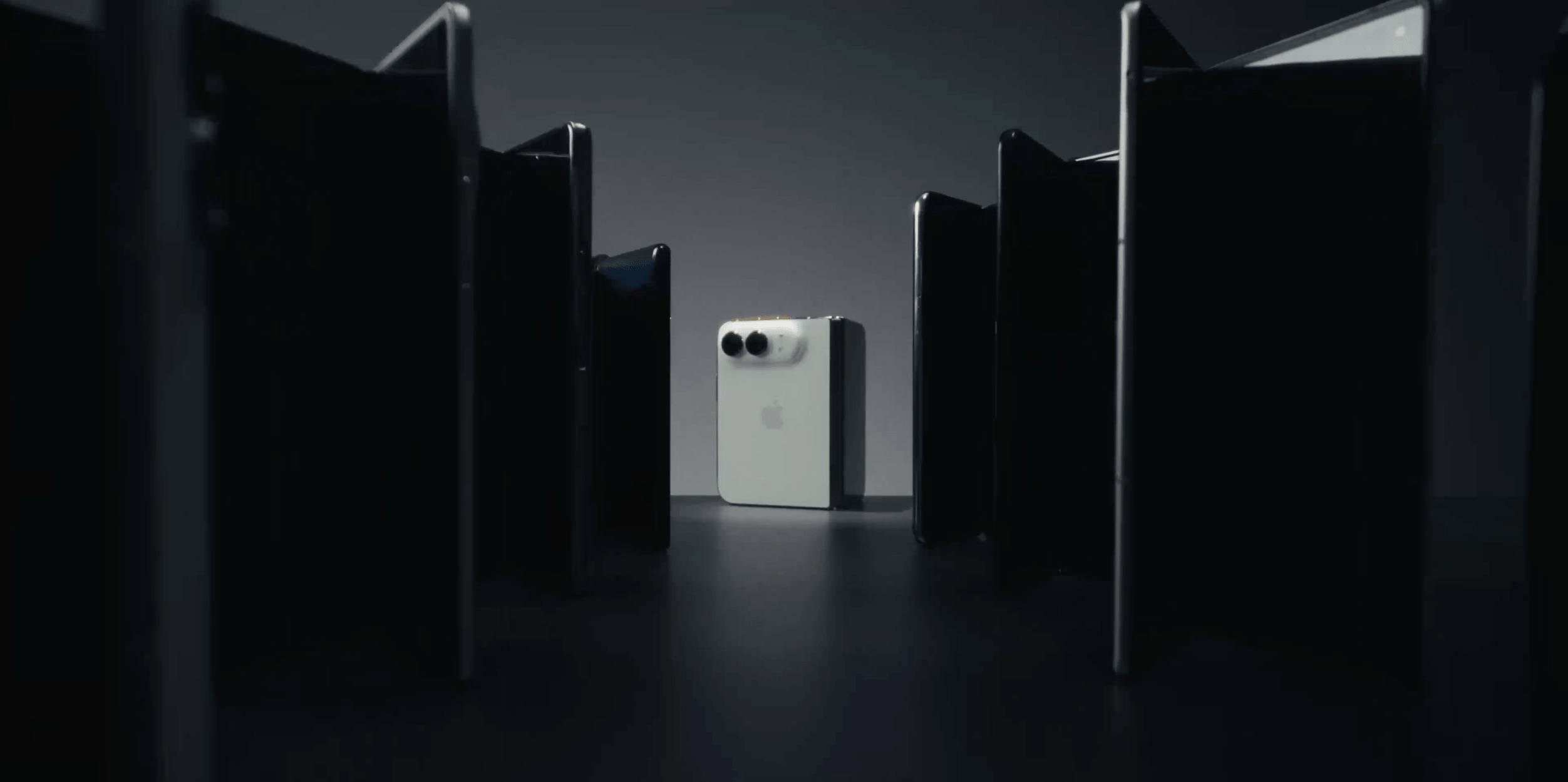 A white smartphone with a dual-camera setup is centrally placed on a dark surface, surrounded by several black smartphones standing upright in a semicircle, all against a dark background.