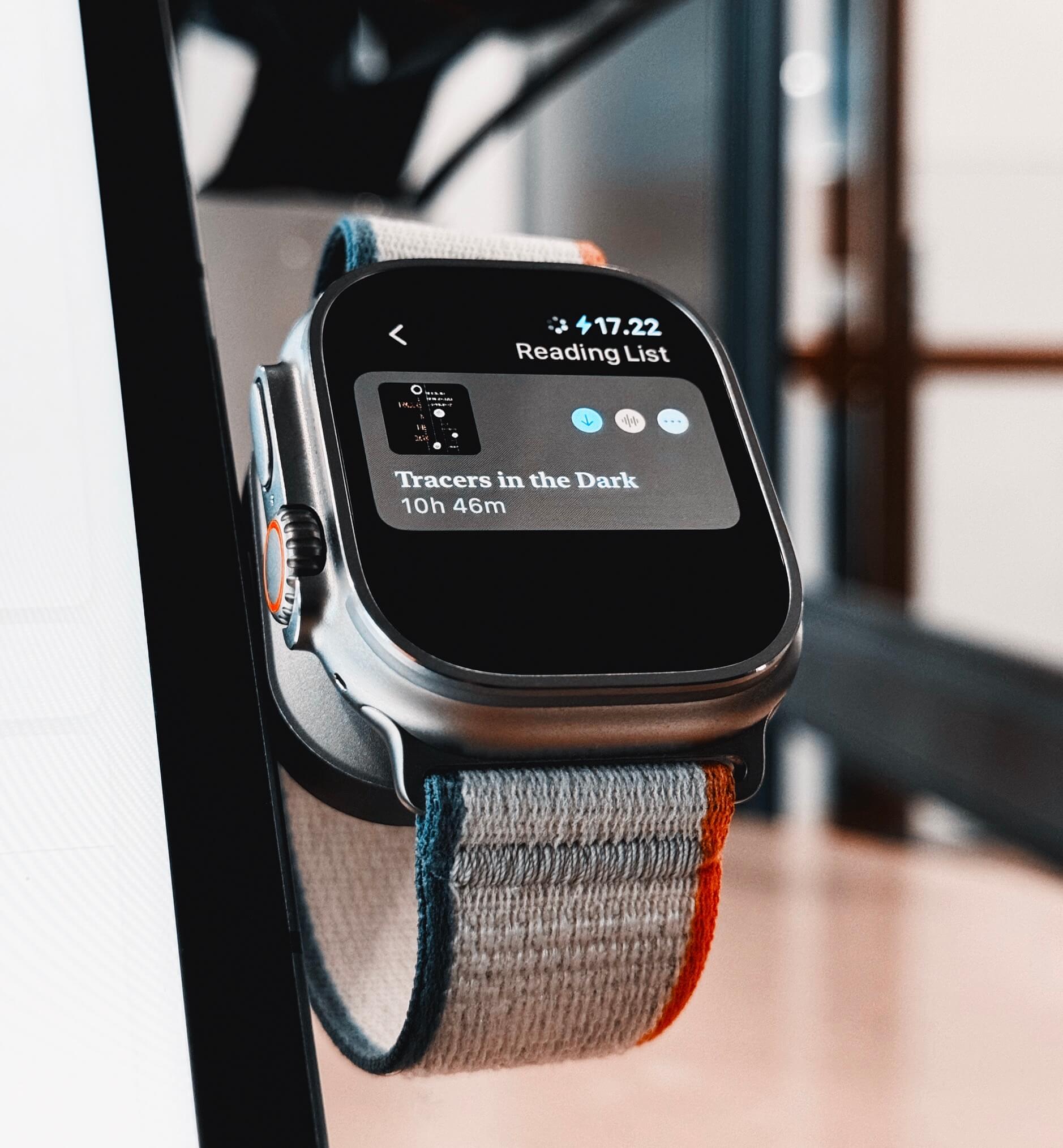 Das ist einer Apple Watch Ultra, die eine Leseliste mit dem Titel "Tracers in the Dark" und einer verbleibenden Lesezeit von 10 Stunden und 46 Minuten anzeigt. Das Uhrarmband hat farbige Streifen in Blau, Grau und Orange.