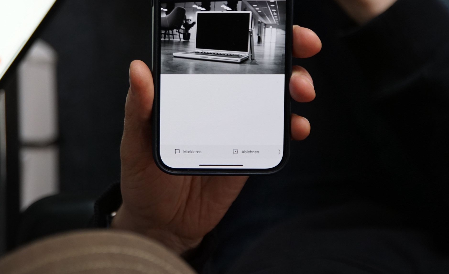 Ein Hand hält ein iPhone, auf dessen Bildschirm eine schwarz-weiße Fotografie eines Laptops zu sehen ist. Unter dem Bild stehen auf Deutsch die Worte "Markieren" und "Ablehnen".