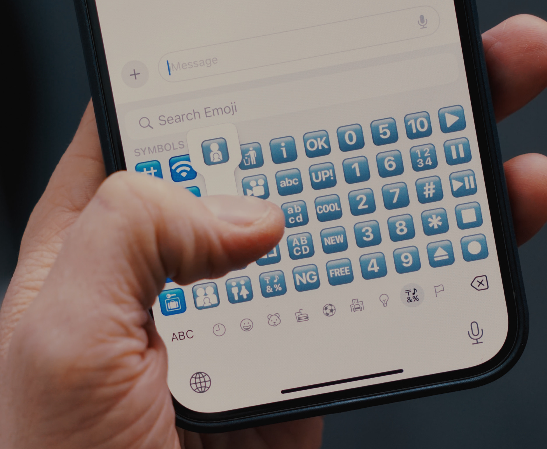 Das Bild zeigt eine Nahaufnahme einer Hand, die ein Smartphone hält, auf dessen Bildschirm eine Emoji-Tastatur zu sehen ist. Der Daumen der Hand ist dabei, ein Emoji auszuwählen.