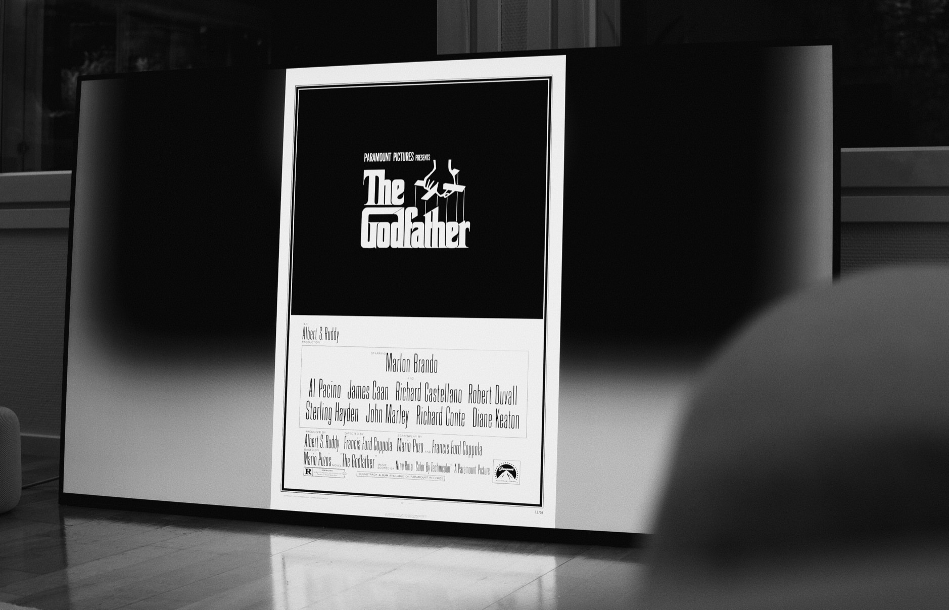 Das Foto zeigt ein Schwarz-Weiß-Bild eines Filmplakats von "Der Pate" mit dem berühmten Puppenspieler-Logo, umgeben von einem weißen Rand, das in einem modernen Raum gegen einen Fernseher lehnt. Auf dem Plakat sind die Namen der Hauptdarsteller und des Regisseurs sowie das Logo von Paramount Pictures zu erkennen.