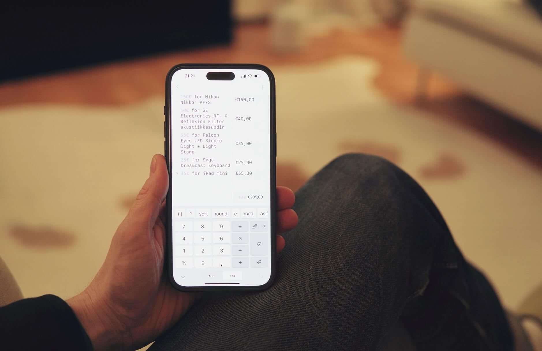 Auf dem Bild ist eine Hand zu sehen, die ein Smartphone hält. Auf dem Bildschirm des Smartphones ist eine Liste von Gegenständen mit ihren Preisen in Euro und ein Taschenrechner zu sehen.