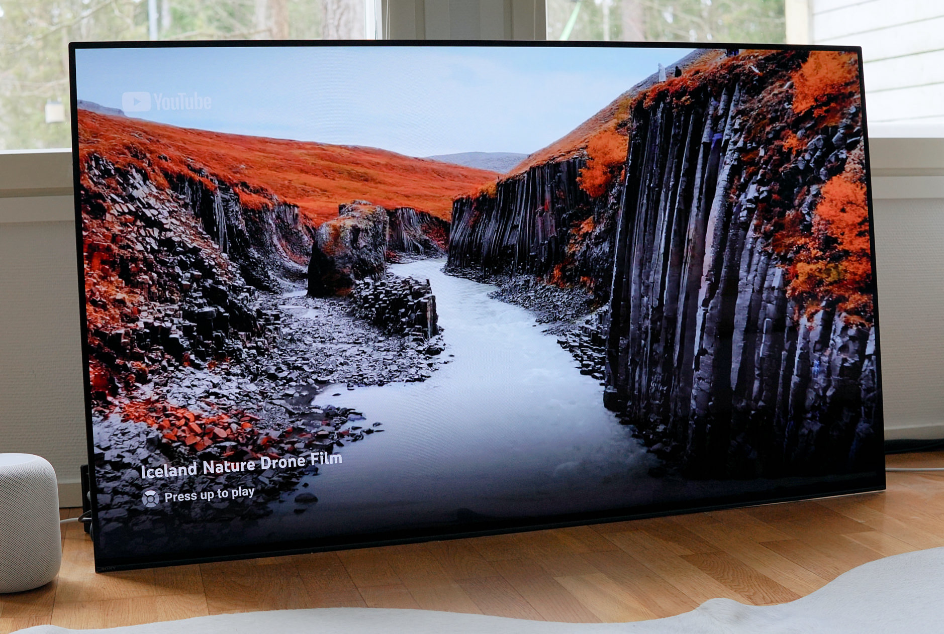 Das Bild zeigt einen großen Fernseher in einem hellen Raum, der ein YouTube-Video mit dem Titel "Iceland Nature Drone Film" anzeigt. Die Szene auf dem Bildschirm stellt eine beeindruckende Landschaft mit einer Schlucht dar, umgeben von roten Moosflächen und basaltischen Felsformationen.