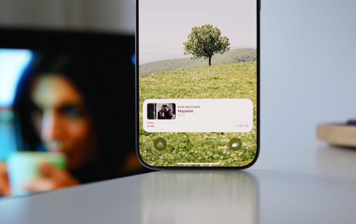Auf diesem Bild ist ein Smartphone zu sehen, das einen Film namens "Haywire" aus dem Jahr 2011 abspielt. Der Bildschirm zeigt im Hintergrund eine idyllische Landschaft mit einem einzelnen Baum auf einer grünen Wiese und sanften Hügeln. Am unteren Rand des Bildschirms sind die Wiedergabezeit von 5:46 und die verbleibende Zeit von -1:27:14 zu sehen, sowie zwei Schaltflächen für Download und Wetter.