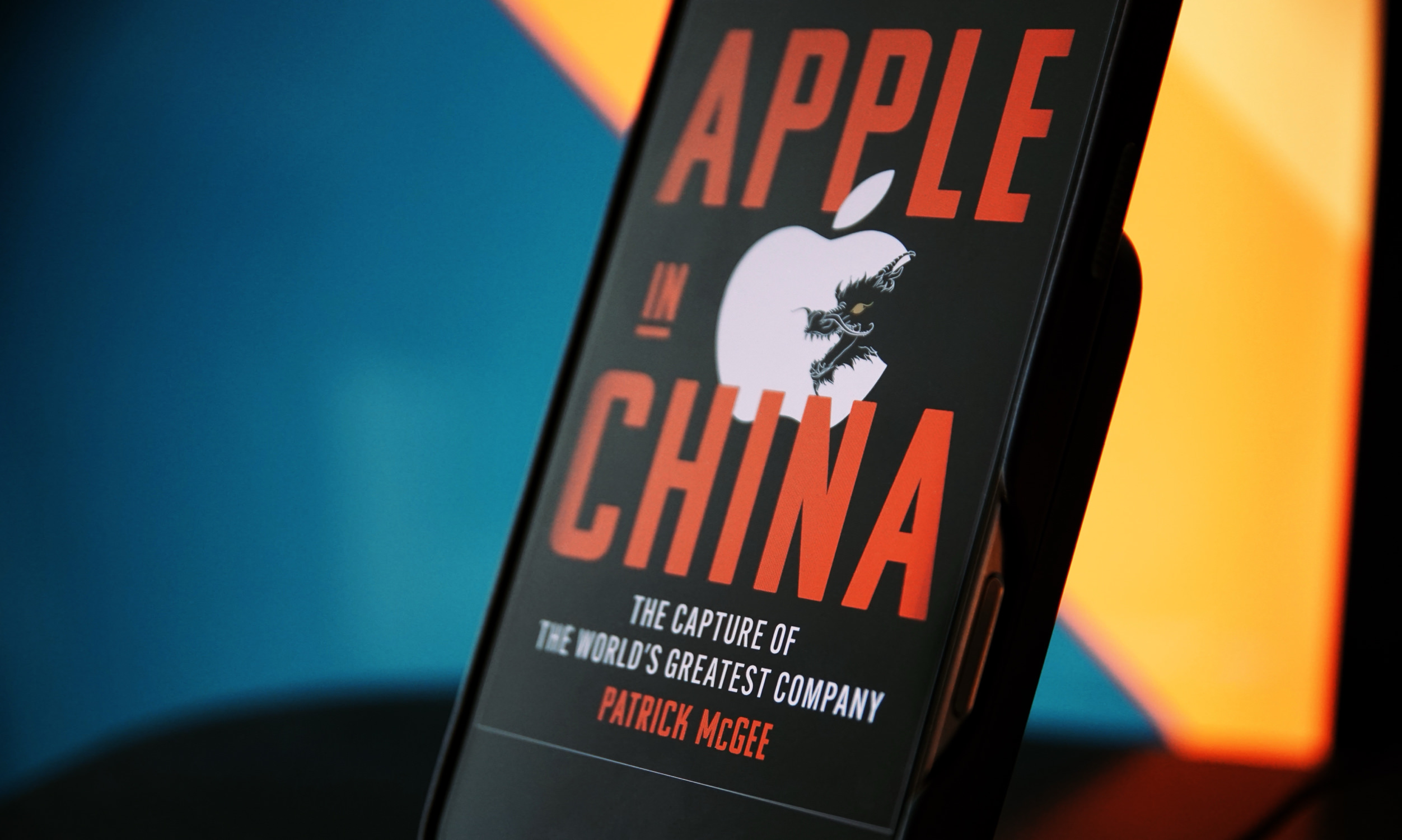 Das Bild zeigt das Buch "Apple in China" von Patrick McGee, das auf einem Ständer präsentiert wird. Der Titel ist auffällig in großer Schrift gehalten, und im Hintergrund ist das bekannte Apple-Logo mit einem Drachen kombiniert. Die Farbgebung ist modern, mit einem Kontrast zwischen Schwarz, Rot und blauen Akzenten.