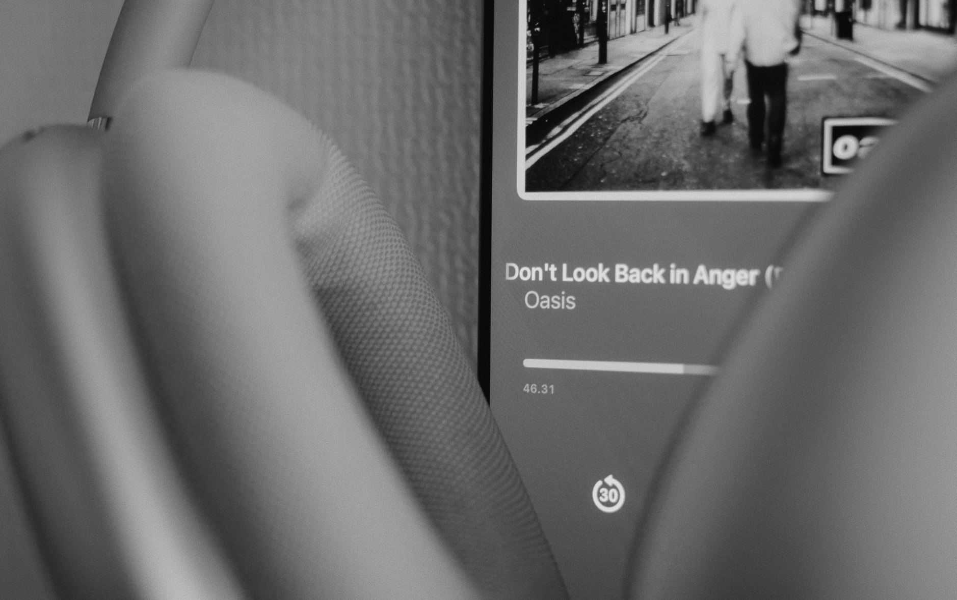 Das Bild zeigt einen Musikplayer auf einem Smartphone-Bildschirm, der den Song "Don't Look Back in Anger" von Oasis abspielt, mit einer Laufzeit von 46:31 Minuten und einem schwarz-weißen Foto einer Person auf einer Straße als Cover. Im Vordergrund ist verschwommen ein Teil eines Kopfhörers oder Lautsprechers zu erkennen, vermutlich ein HomePod Mini oder ähnliches Audiogerät. Die Aufnahme ist in Schwarz-Weiß gehalten, was dem Bild eine minimalistische und künstlerische Atmosphäre verleiht.