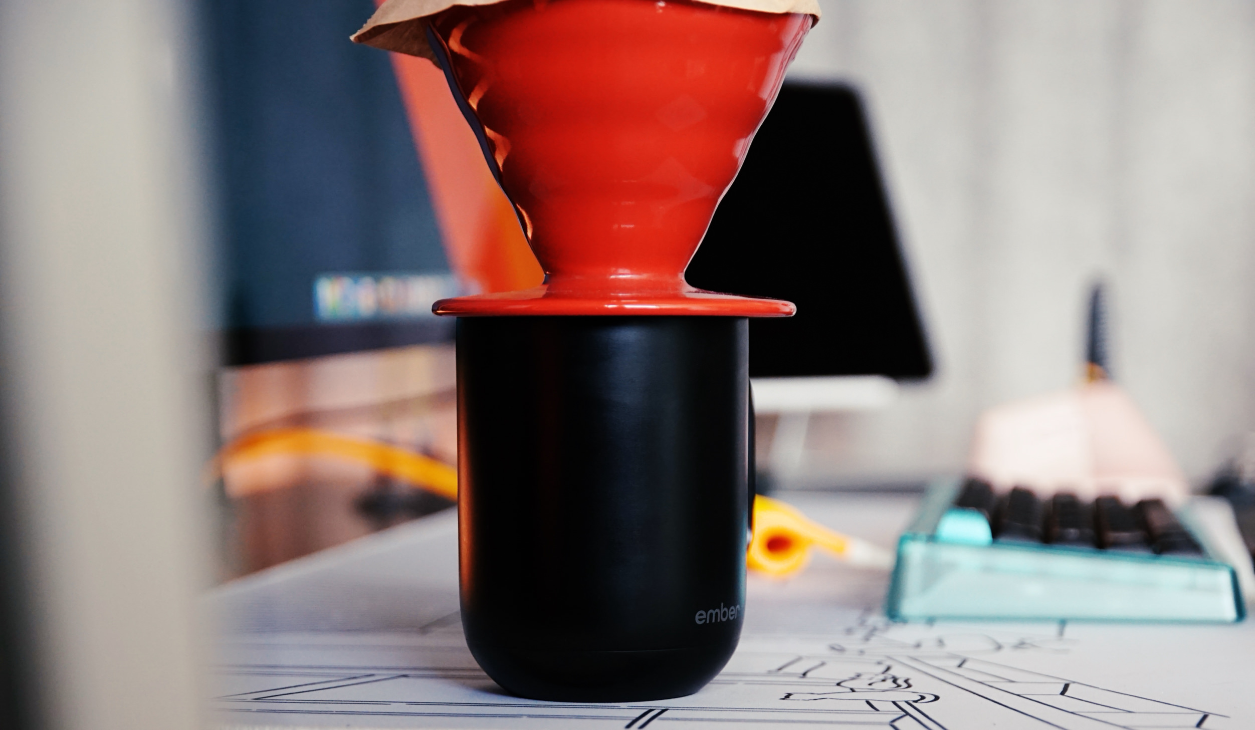 Das Bild zeigt einen roten Kaffeefilterhalter, der auf einem schwarzen Behälter steht. Im Hintergrund sind verschwommen ein Computer und ein Schreibtisch mit Zubehör zu sehen. Die Szene vermittelt eine gemütliche, kreative Atmosphäre zum Kaffeebrauern.