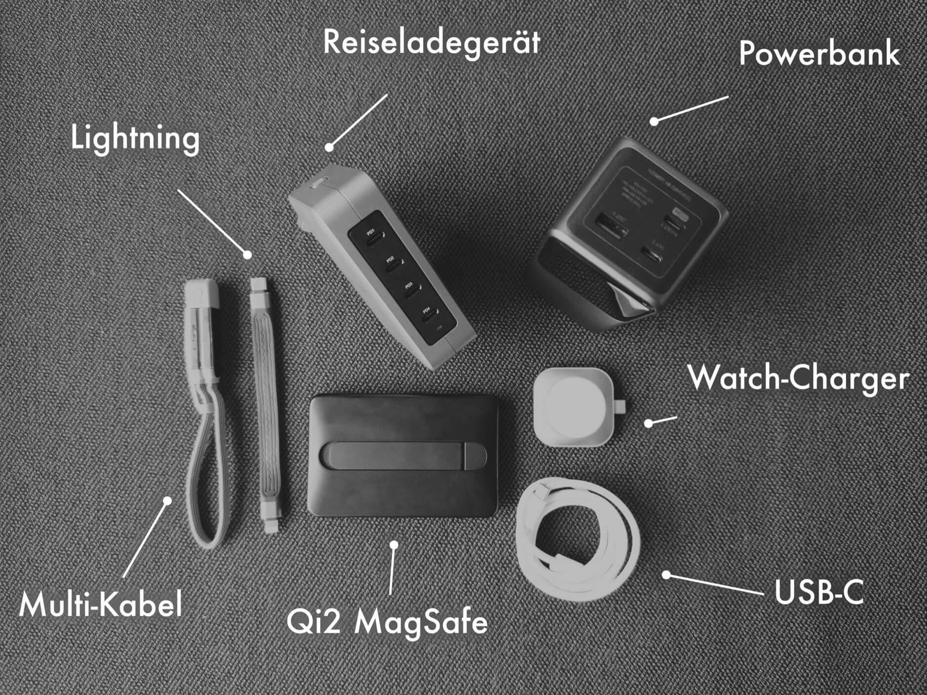 Overhead shot of various electronic charging devices laid out on a gray textile surface, including a travel charger labeled "Reiseladegerät," a power bank, a Qi2 MagSafe charging pad, a watch charger, a multi-cable, a lightning cable, and a USB-C cable.
