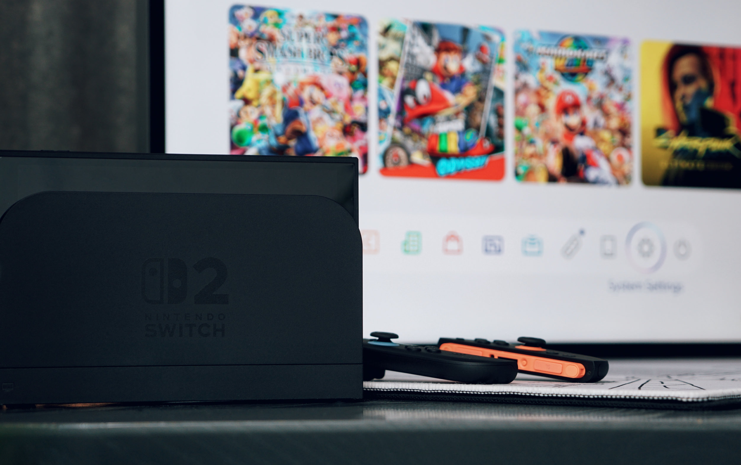 A Nintendo Switch console in its dock, with Joy-Con controllers resting below; on a screen behind the console are game icons and menu options, indicating the console is active.