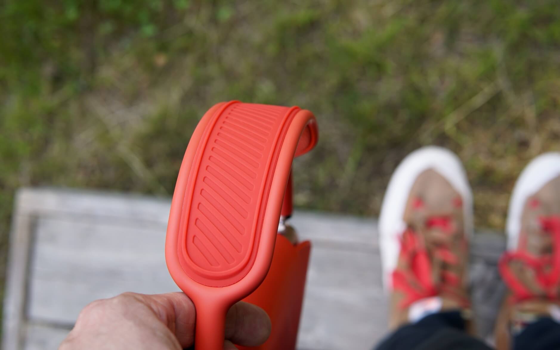 Das Bild zeigt eine Hand, die einen orangefarbenen Silikongriff hält. Im Hintergrund sind verschwommen ein paar Schuhe mit roten Schnürsenkeln zu sehen. Der Untergrund besteht aus Gras und einer Holzoberfläche.