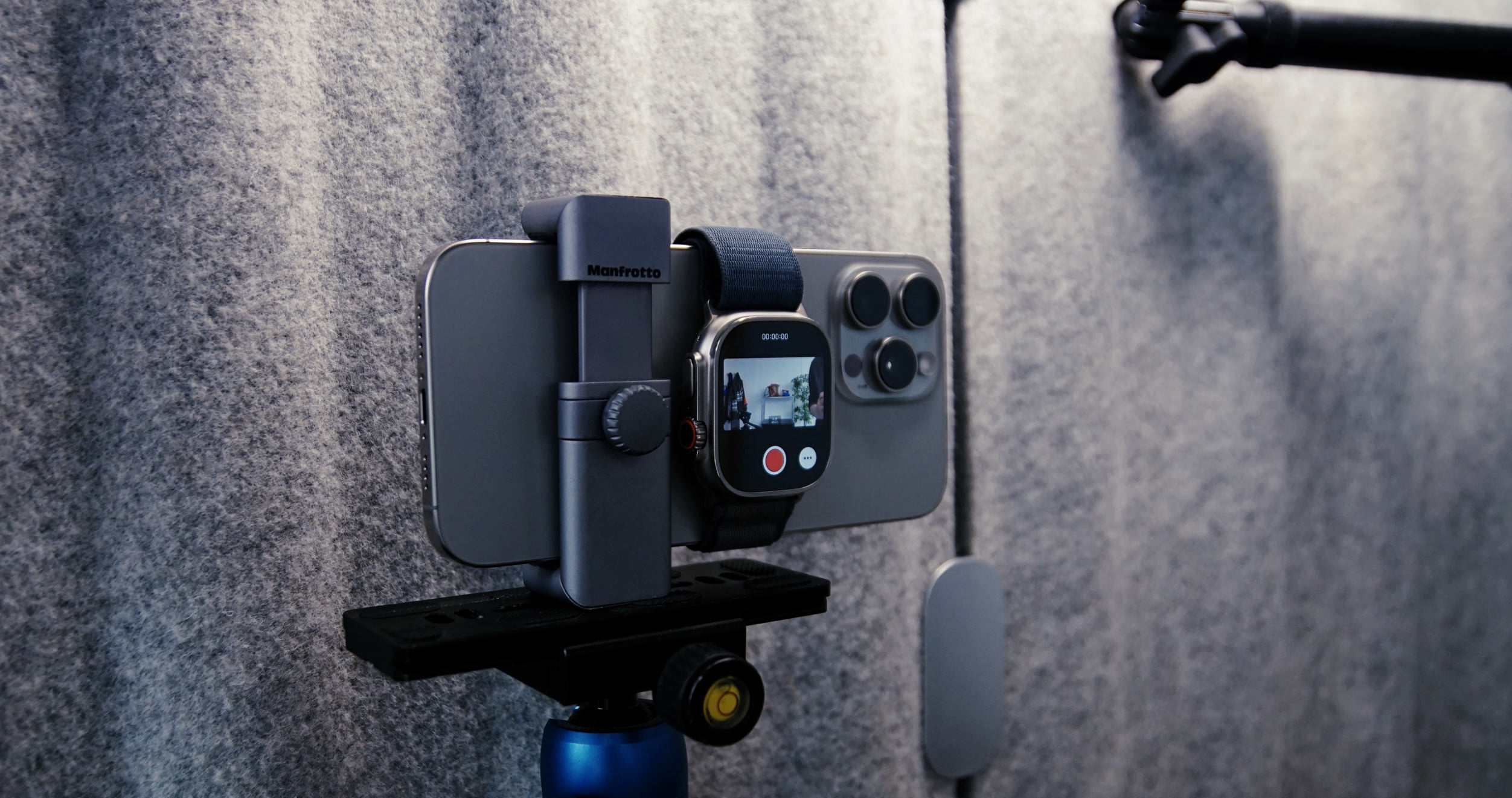 Das Bild zeigt ein Smartphone, das auf einem Stativ montiert ist, zusammen mit einer Smartwatch, die offenbar zur Aufnahme verwendet wird. Im Hintergrund ist eine graue Wand zu sehen, die aus einem strukturierten Material besteht. Diese Anordnung deutet auf eine professionelle Videoaufnahme oder Live-Streaming-Situation hin.