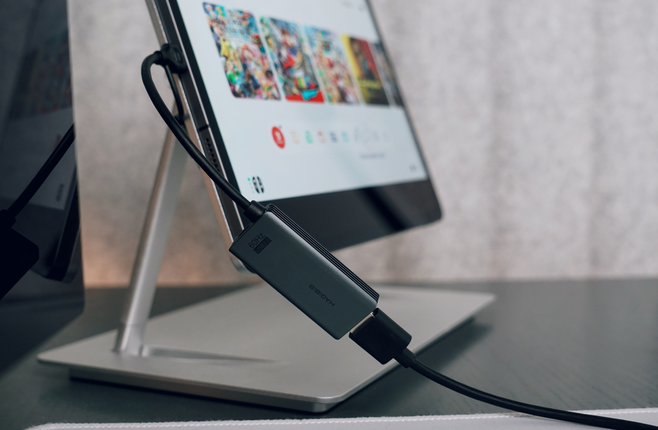 Close-up of a tablet displaying a gaming interface, connected by a black cable to a gray Hagibis USB 3.0 HDMI adapter. The tablet is propped on a silver stand on a dark surface.