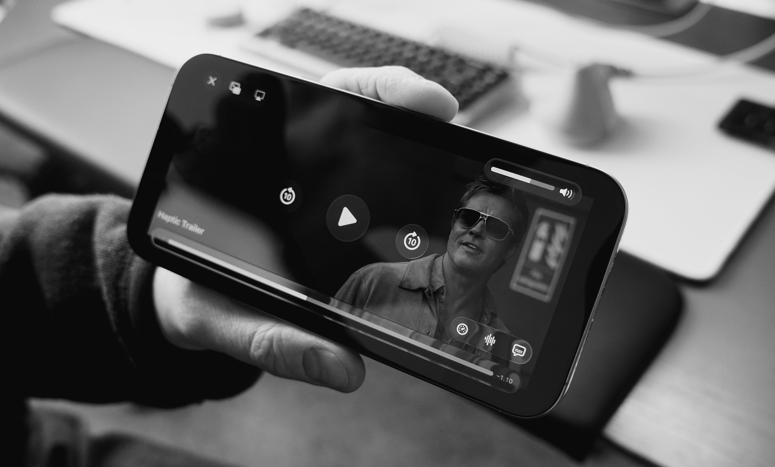 Auf dem Bild hält eine Person ein Smartphone in der Hand, auf dem ein haptischer Trailer abgespielt wird. Der Bildschirm zeigt einen Mann in Sonnenbrille, der aus einem Film oder Video stammt. Im Hintergrund sind einige unscharfe Büroobjekte zu erkennen.