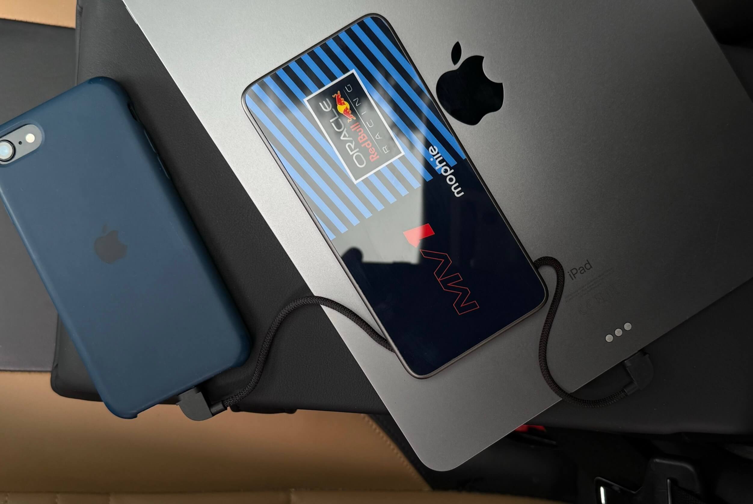A modern mobile device with a Red Bull Racing design case and a connecting cord lies across an open grey Apple iPad, adjacent to a dark blue iPhone with a connected charging cable.