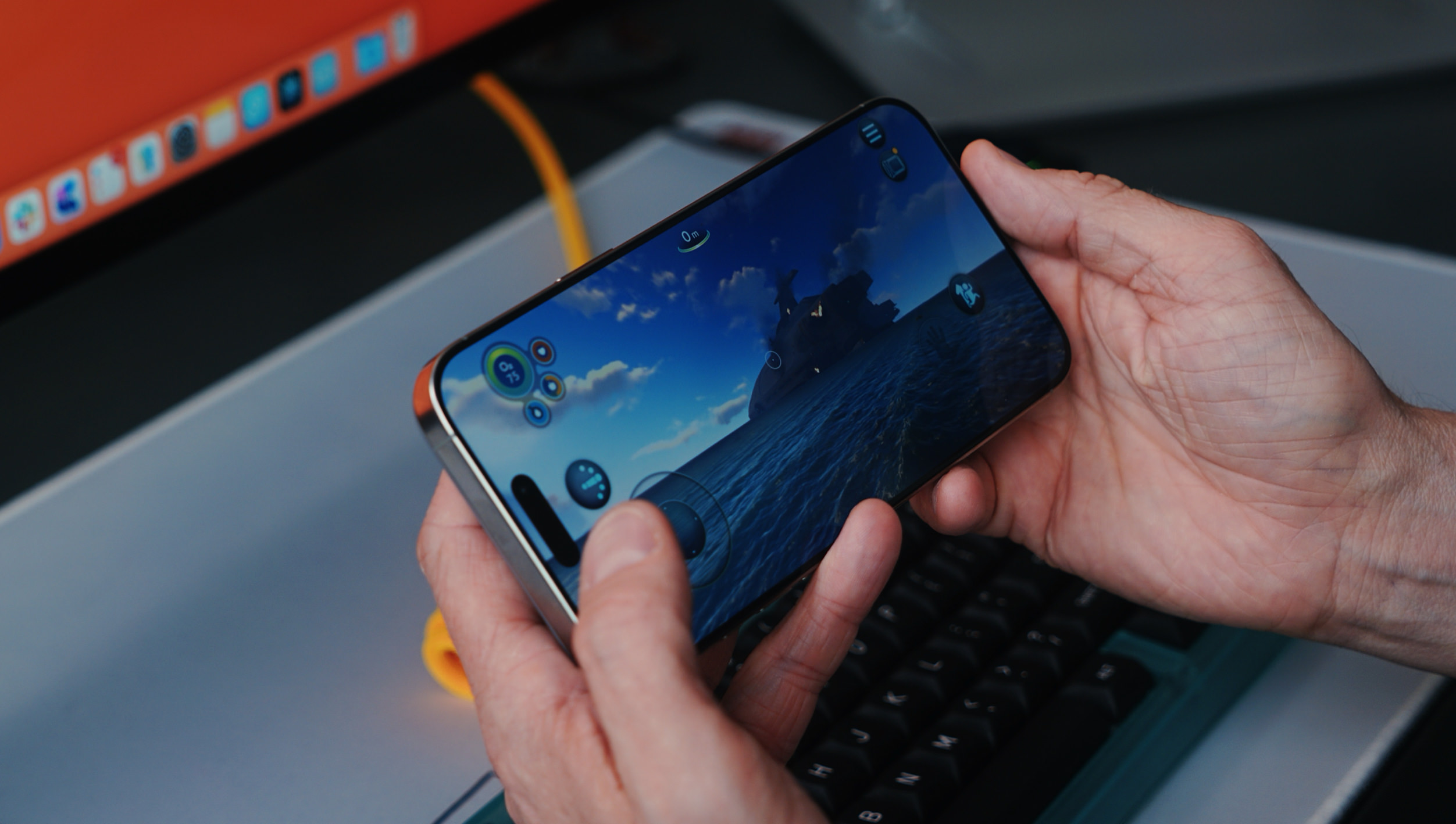 Person holding a smartphone with a game displayed, showing a blue ocean scene with islands, skies, and a submarine. The phone's screen displays virtual controls for gameplay. The background includes a keyboard, and an orange computer screen is visible in the background.