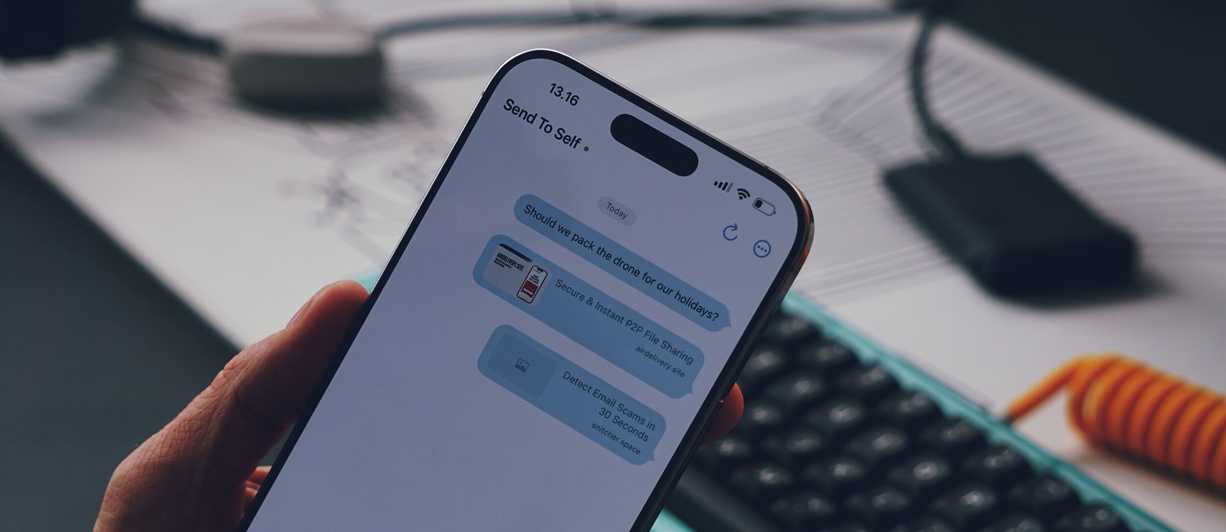 A hand holding a smartphone displaying a messaging app conversation with "Send To Self." The conversation includes three blue message bubbles: "Should we pack the drone for our holidays?," "Secure & Instant P2P File Sharing, airdelivery.site," and "Detect Email Scams in 30 Seconds, snitcher.space." The time 13:16 is shown at the top, along with the phone's status bar. The background shows a partially visible desk, a keyboard, and some cables.