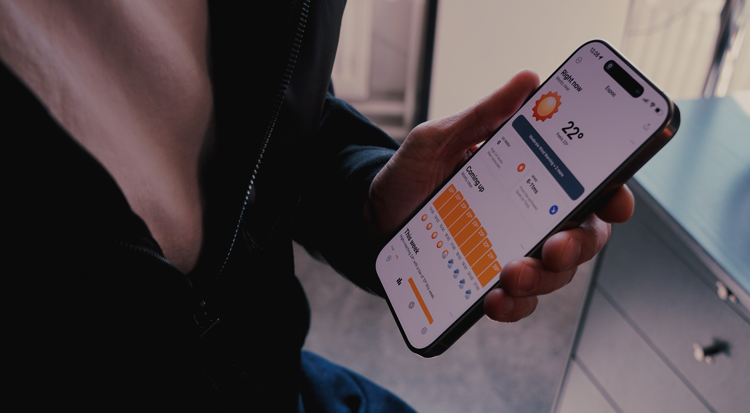 A person's hand holding a smartphone displaying a weather forecast for Espoo. The screen shows a sunny icon, a temperature of 22 degrees Celsius, and a 'Moderate Wind Warning'. The weather forecast includes 'Coming up' hourly details and a 'This week' overview. The person is wearing a dark jacket, with the zipper in focus.