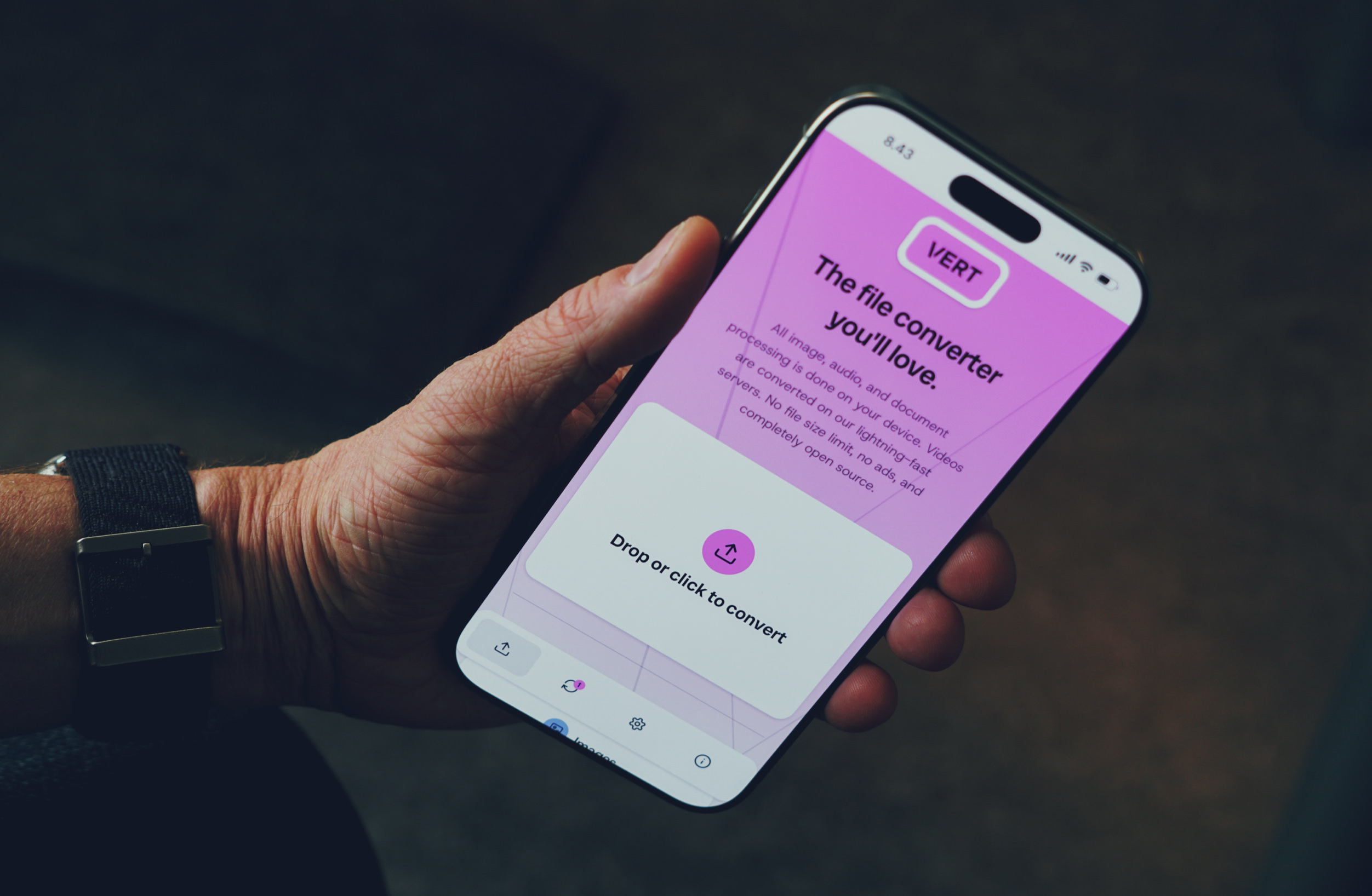 A hand holds a smartphone displaying the "VERT" file converter app interface, with a pink UI, upload button, and text promoting its image, audio, and document conversion capabilities, highlighting device-based processing and no file size limits.
