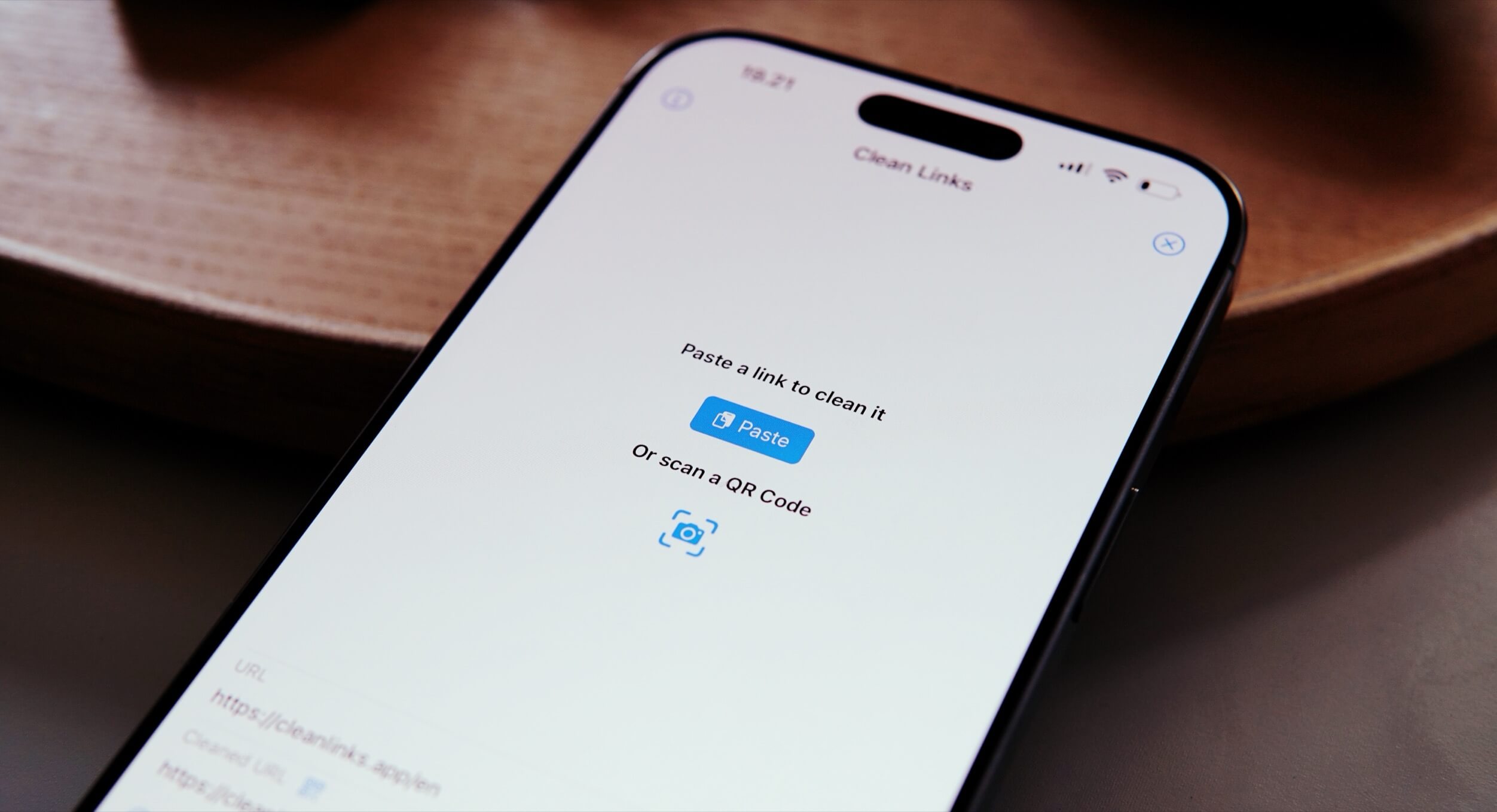 Close-up of an iPhone screen displaying the "Clean Links" app interface. The app prompts the user to paste a link, offering a "Paste" button, or to scan a QR code. The top of the screen shows the time, signal strength, and battery icon. The phone rests on a wooden surface.