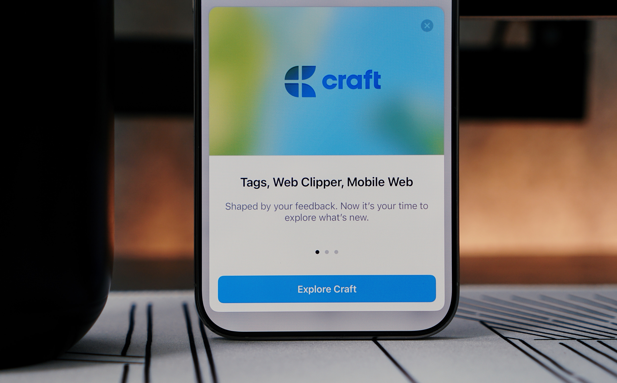A smartphone displaying the Craft app welcome screen. The screen has a blurred blue and green background with the Craft logo at the top, and the text "Tags, Web Clipper, Mobile Web" followed by a description and an "Explore Craft" button. The phone sits on a patterned surface next to a black object, likely a cup.