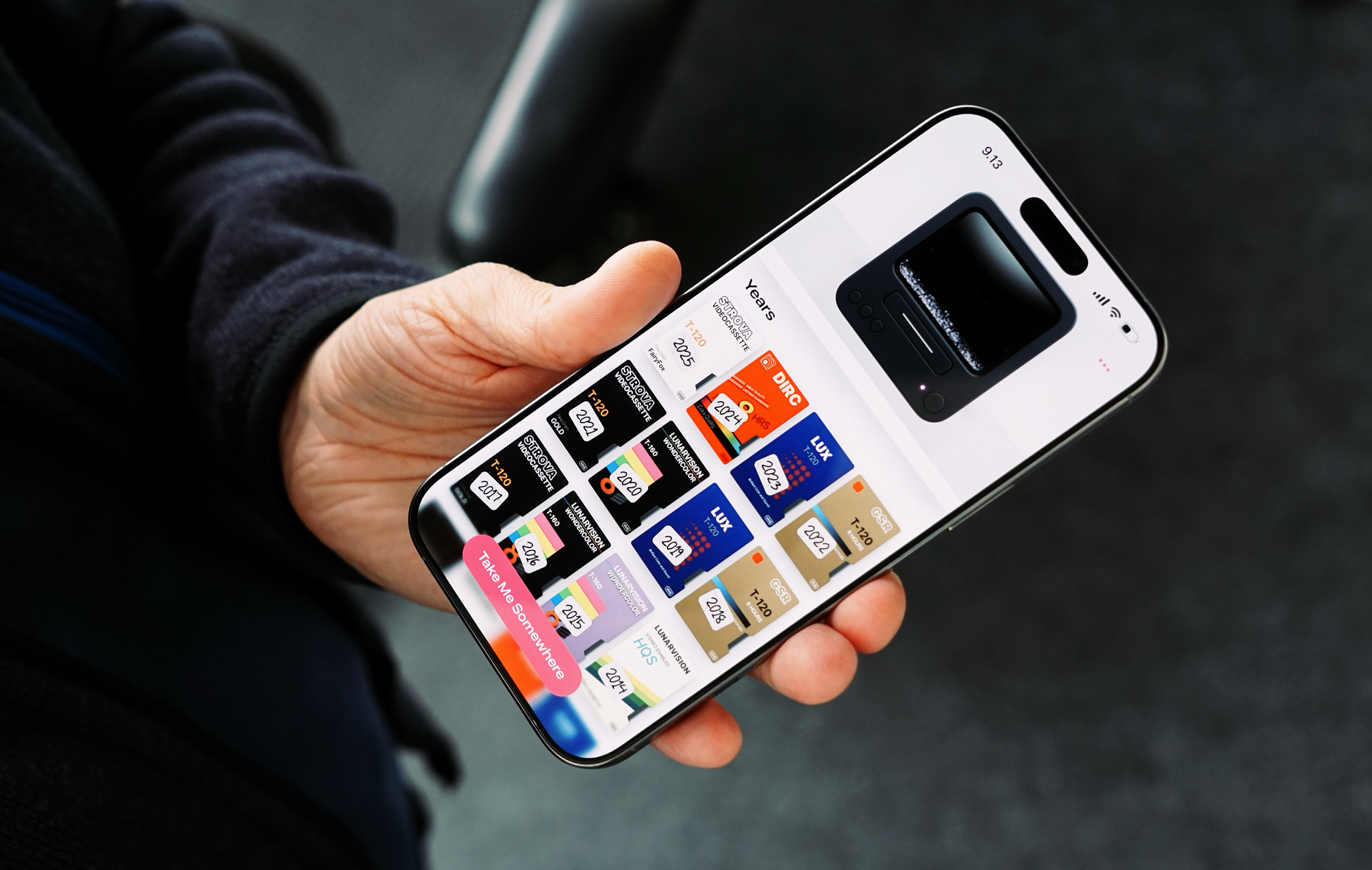 A hand holding an iPhone displaying a user interface with the title "Years" and various blocks representing different years, each block featuring text related to video cassette tapes, the interface includes a "Take Me Somewhere" call to action.