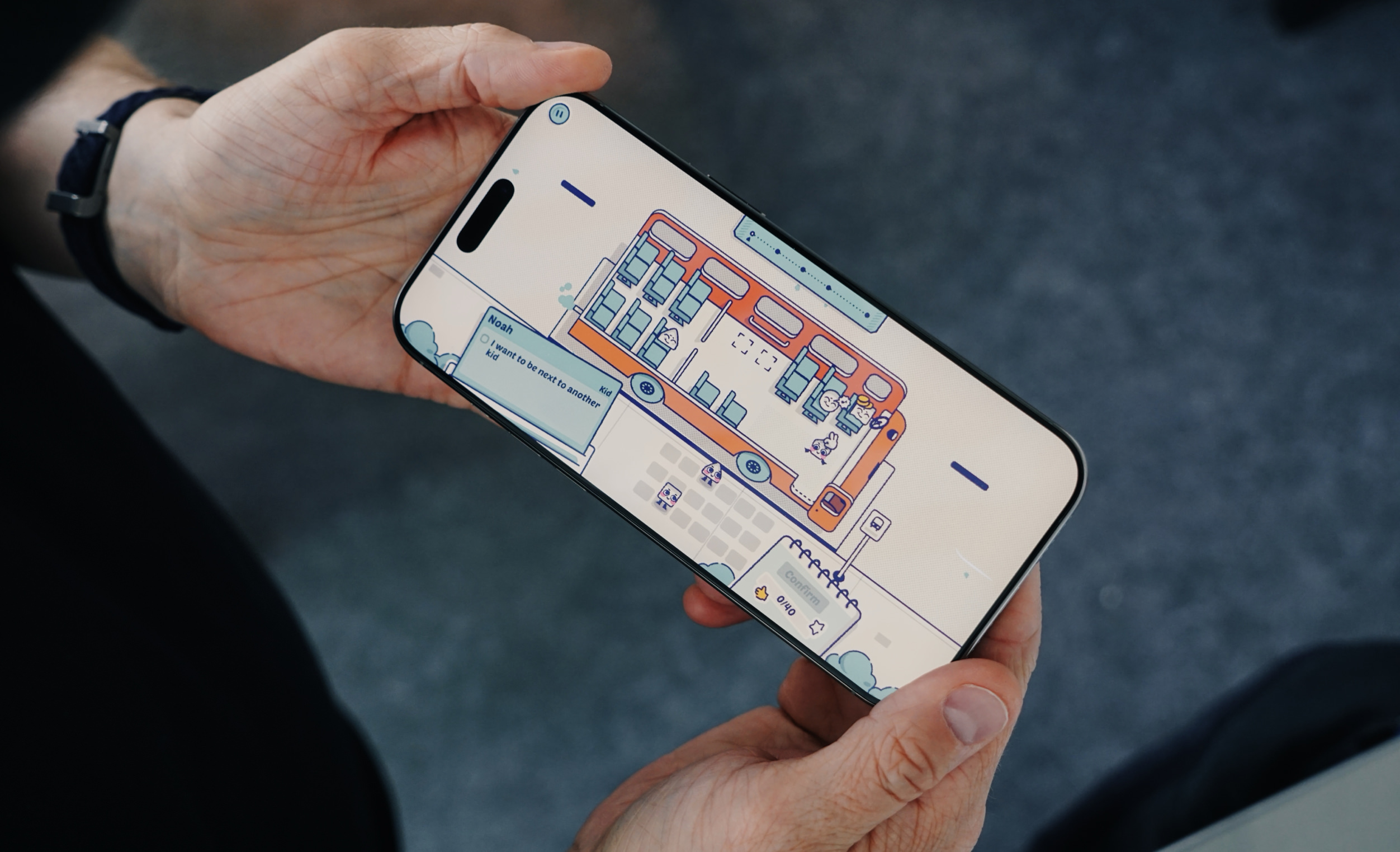 A smartphone held by hands displays a cartoon bus interior with characters, likely a mobile game. The screen highlights a seating arrangement interface with dialogue bubble "I want to be next to another kid" along with confirmation and currency options.

