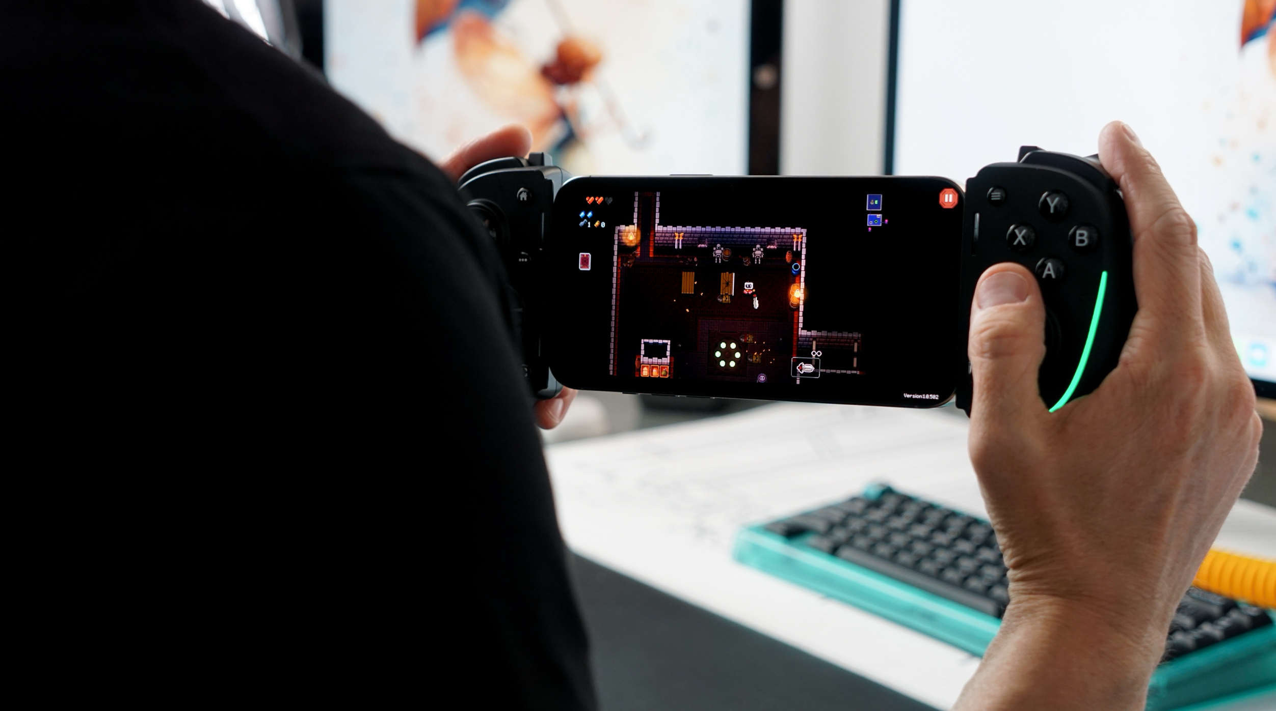 A person holding a smartphone with a gaming controller attached, displaying a top-down view of a pixelated action game, likely a rogue-like, in a dark room setting with characters visible, controller buttons, and game interface elements are on the phone's screen. The hands hold the controller, with the right controller side showing glowing green lights and the buttons. The person is in front of a computer setup, with a keyboard and monitors visible in the blurred background.
