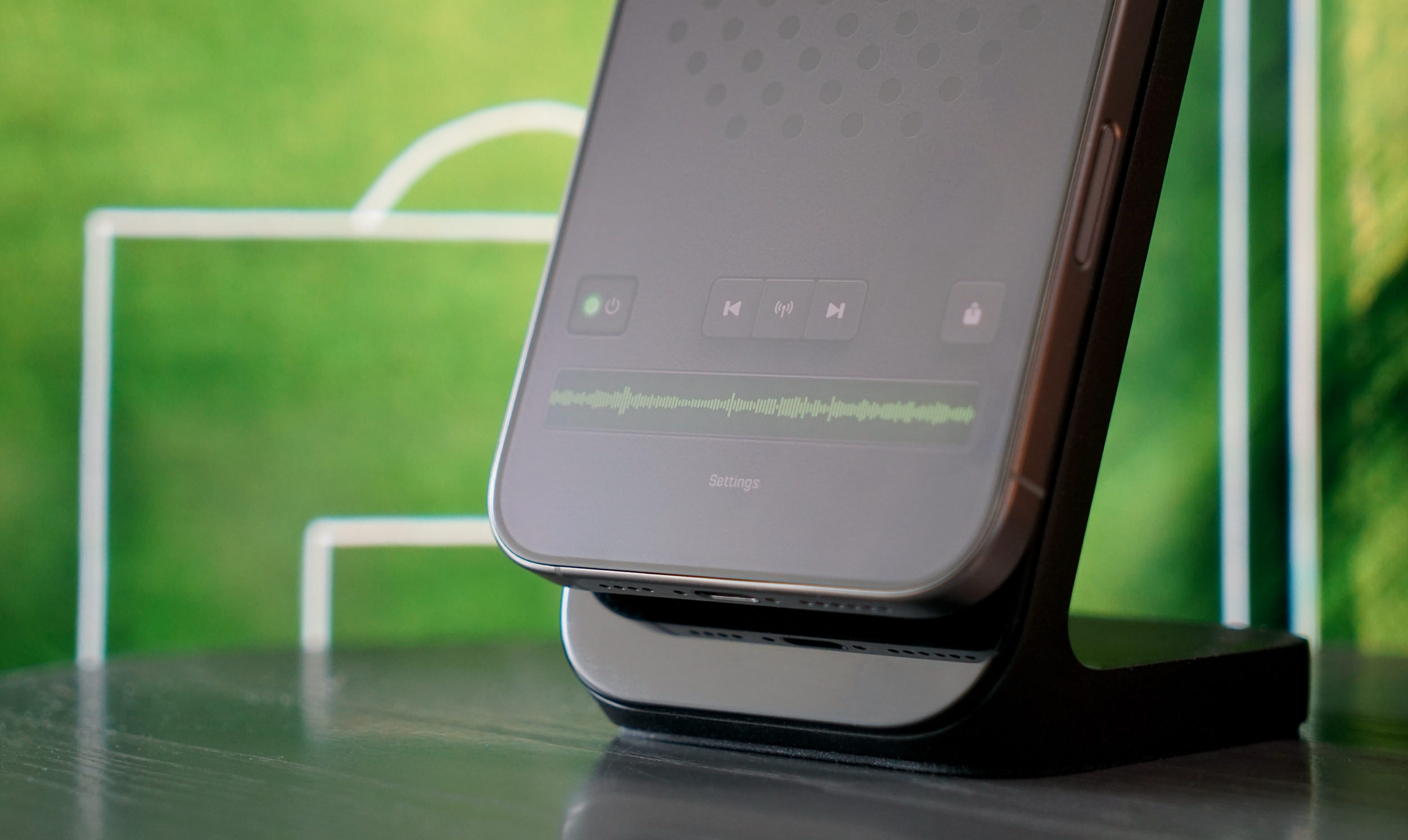 Close-up of a smartphone on a charging stand displaying a music waveform and controls, set against a blurred green background reminiscent of a soccer field.