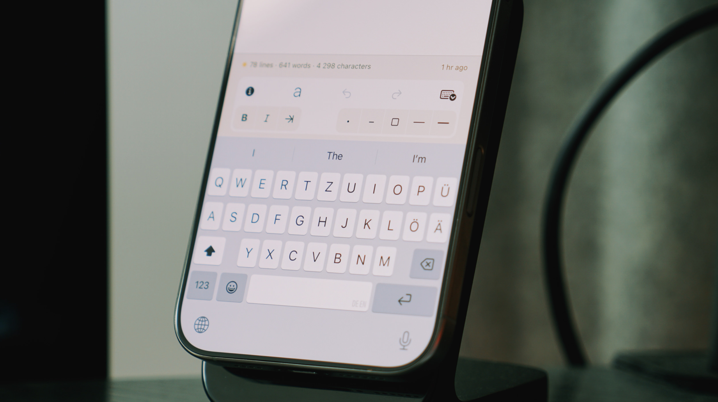 Close-up of a smartphone screen displaying a writing interface with a keyboard, likely a document, resting on a black stand. The screen's interface shows text formatting options above the keyboard, with the letters "QWERTY..." visible. The screen displays the words "The" and "I'm" as context for the text.