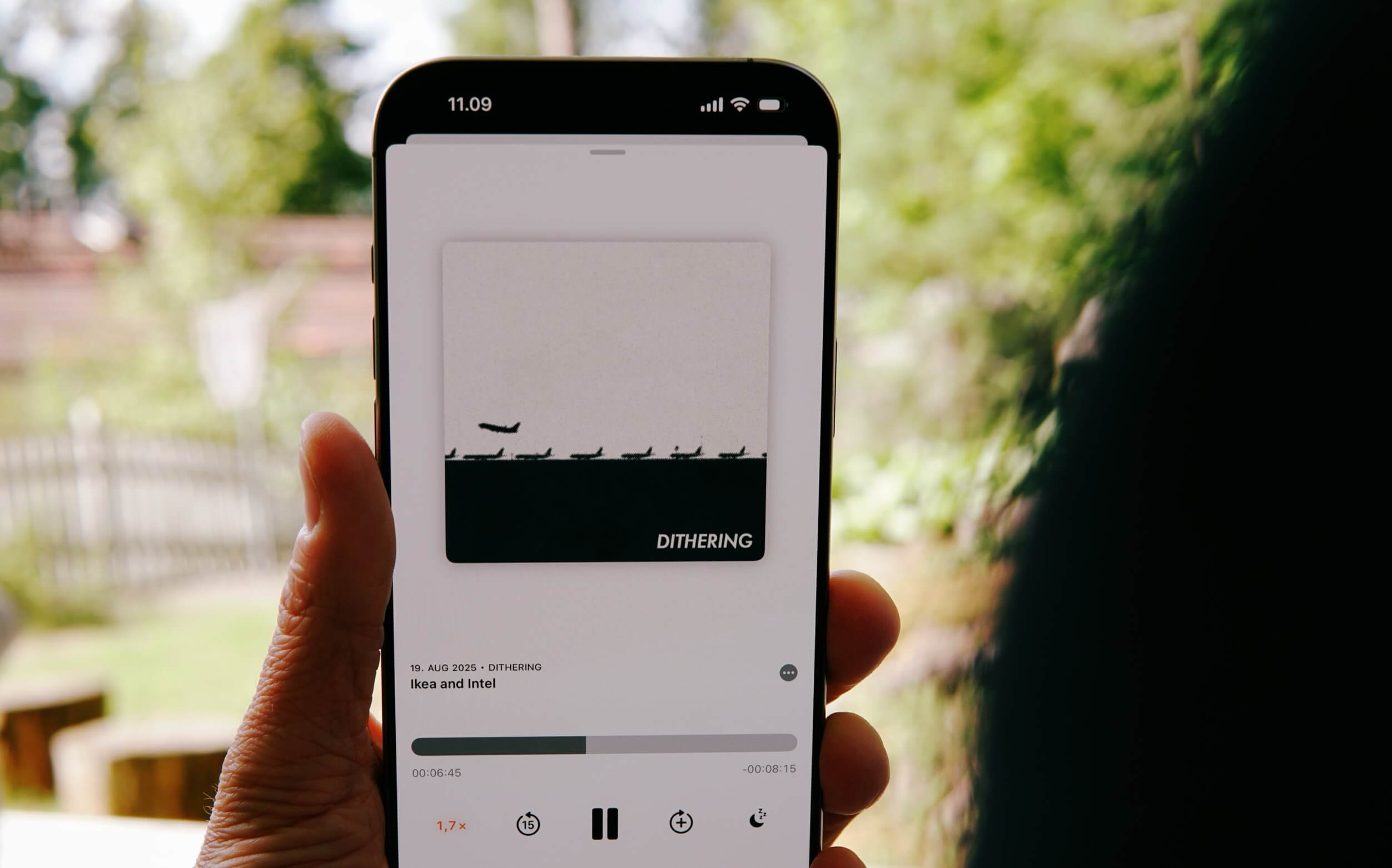 A hand holding a smartphone displaying a podcast titled "Dithering" with a thumbnail of airplanes on a black stripe. The podcast is from August 19, 2025, about Ikea and Intel, with playback controls and time displayed.
