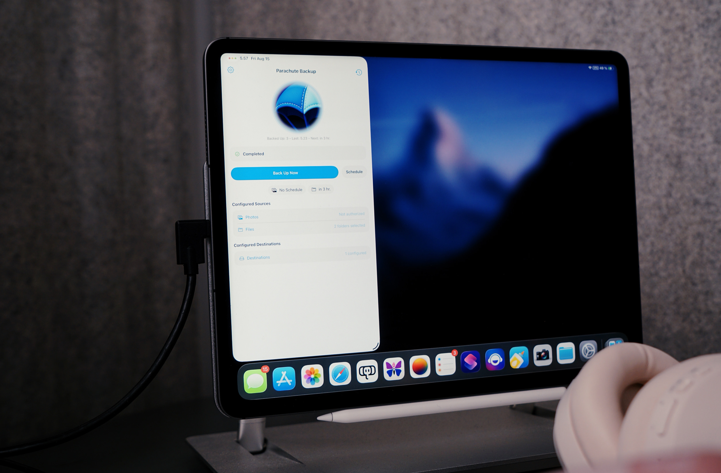 A close-up of an iPad displaying the "Parachute Backup" app with a progress overview. The screen shows the backup status as "completed," with options to "Back Up Now" or "Schedule". The app details the backup configuration, including sources like "Photos" (not authorized) and "Files", and destinations. The iPad is connected to a power cord and is placed on a stand. On the bottom of the screen, there are several app icons, and there's also a white pencil. In the lower right corner are white headphones partially visible.
