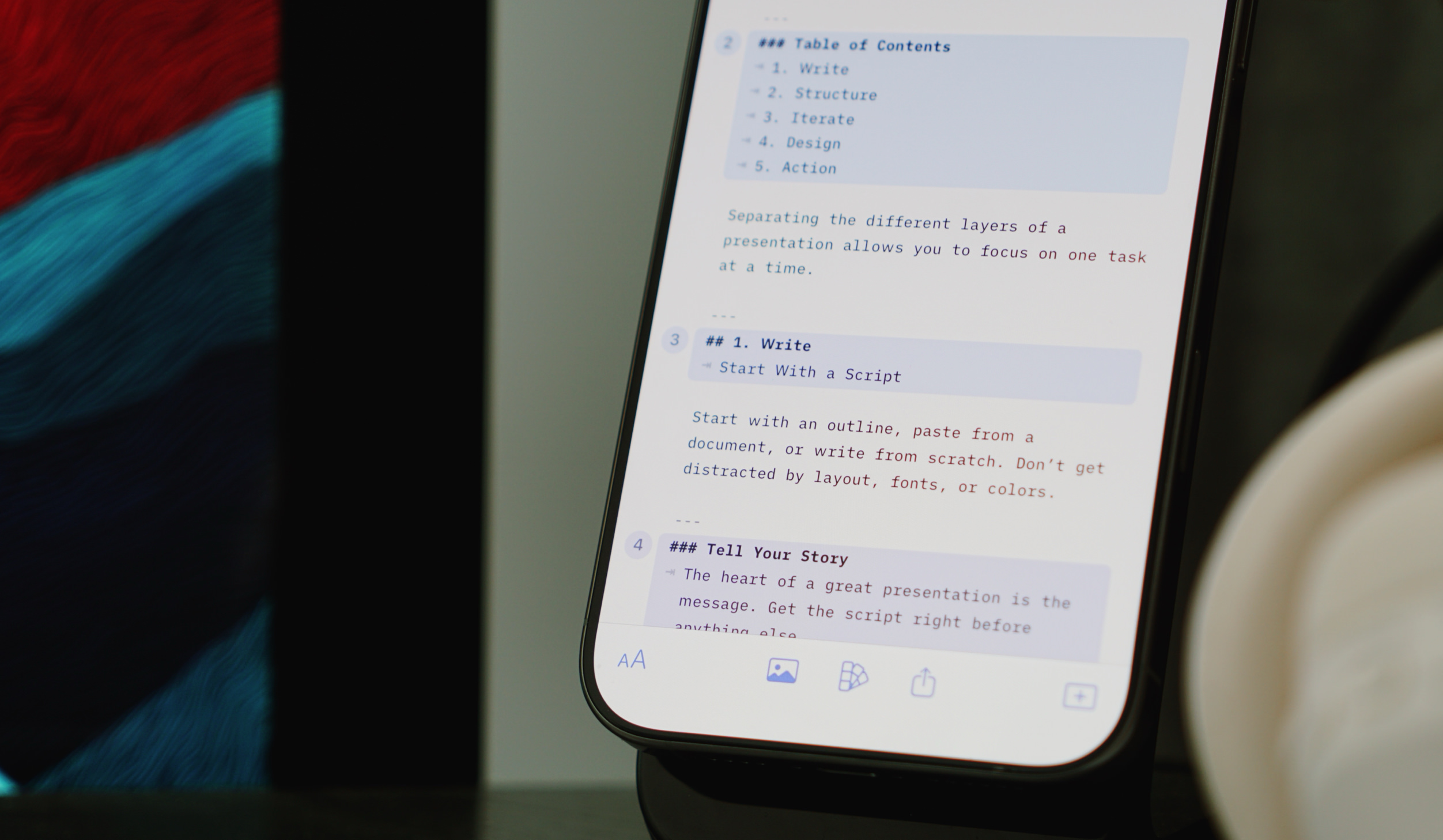Close-up shot of a smartphone displaying a presentation outline with numbered sections on writing a presentation, with a table of contents including Write, Structure, Iterate, Design, and Action. The screen shows content related to scripting a presentation. The phone is set against a blurred background with abstract red and blue color patterns and a white cup on the right.
