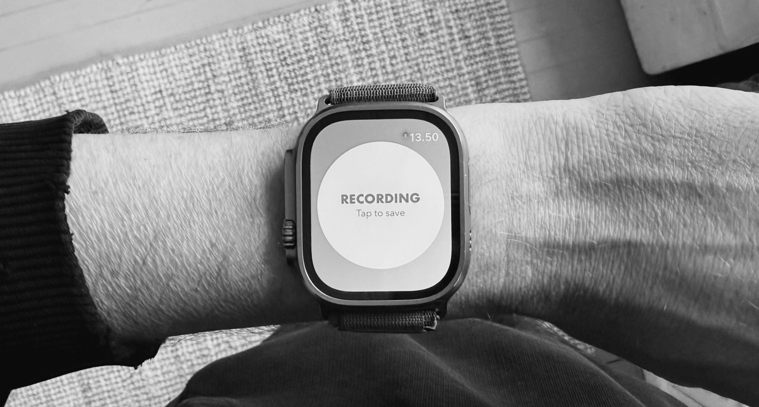Close-up, black and white image of a wrist wearing a smart watch with a screen displaying a "Recording" message. The watch, likely an Apple Watch, is positioned on the center of the wrist. The screen displays the word "RECORDING" and "Tap to save". Above, the time is displayed: 13.50. The band is a light grey nylon fabric. The background shows portions of an arm and a rug or carpet with a textured pattern.
