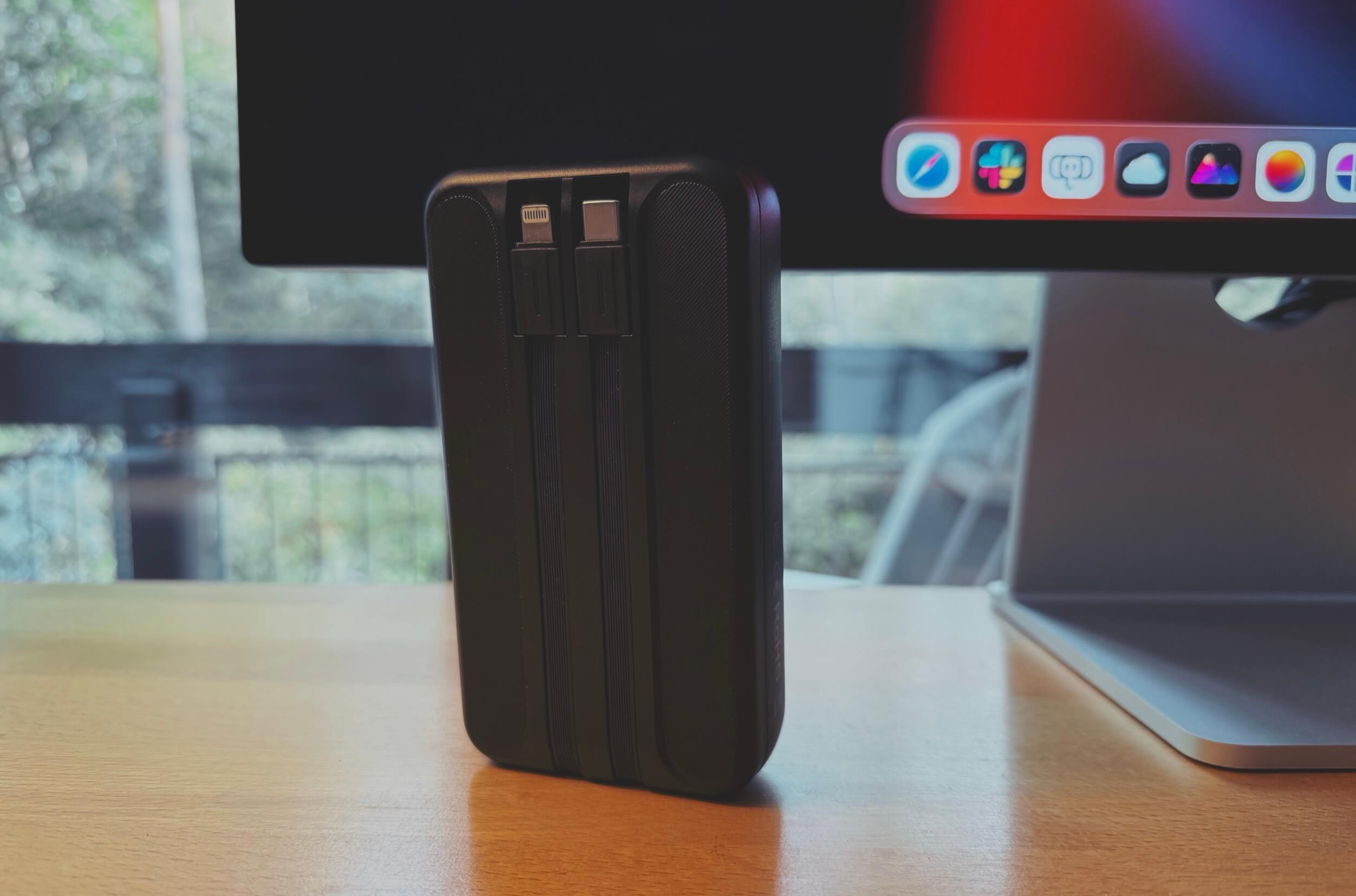 Black portable charger with attached Lightning and USB-C cables, resting on a wooden table in front of a computer monitor displaying a macOS desktop.