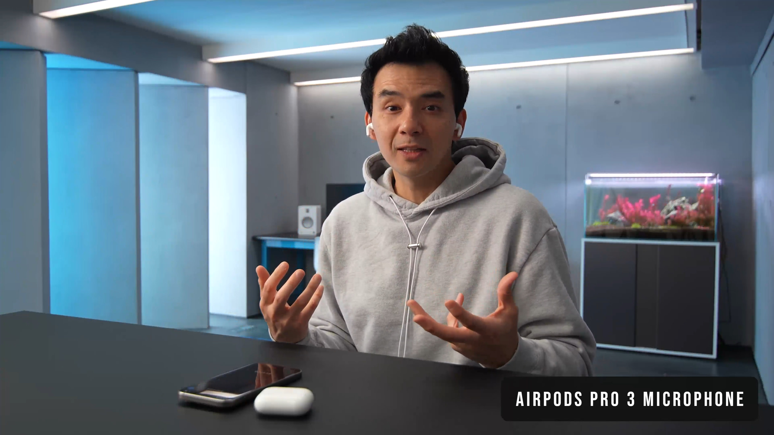 A man wearing AirPods speaks into a microphone while reviewing the Apple AirPods Pro 3, gesturing with his hands at a black table with a smartphone and charging case; a fish tank and studio background are visible.