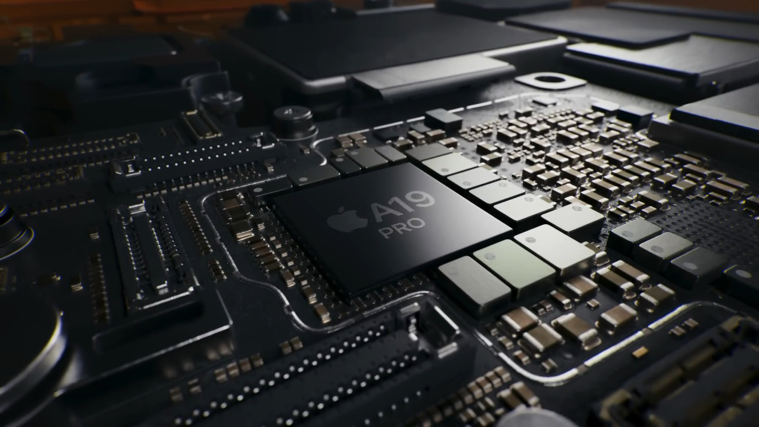 Close-up shot of a computer processor, the Apple A19 Pro chip, centered on a dark circuit board with various micro components and traces visible.