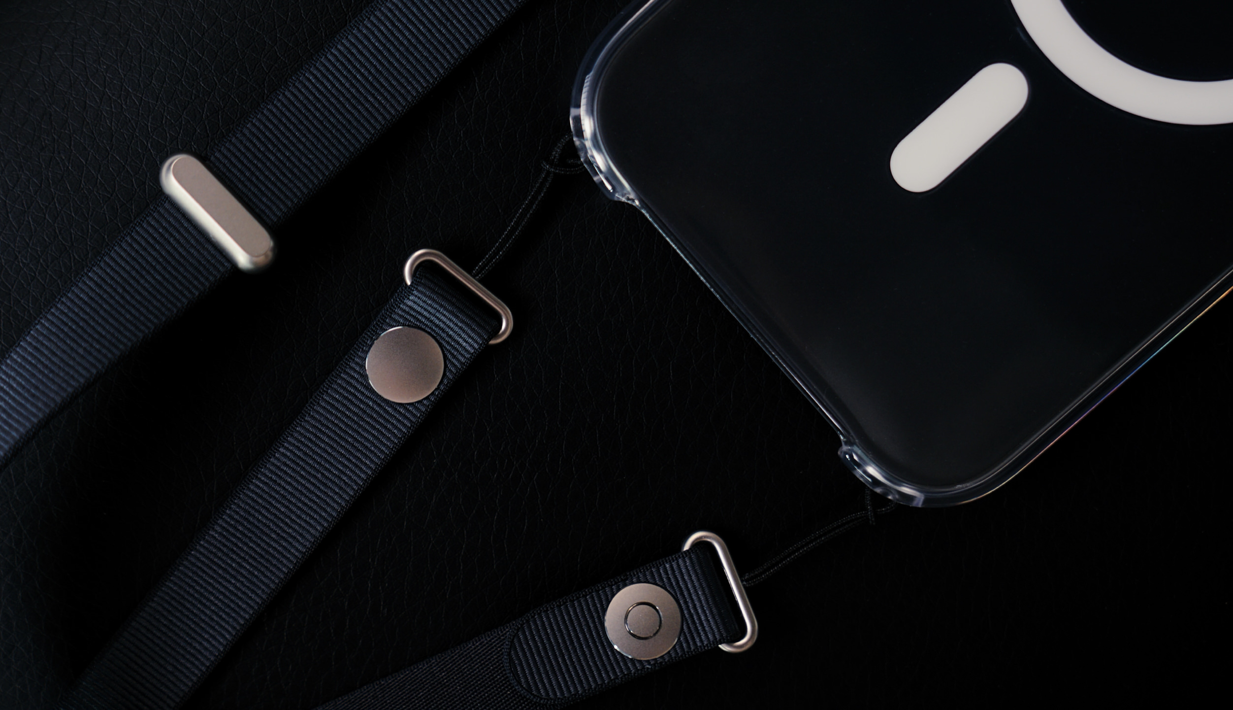 Close-up image of a clear phone case with an attached lanyard and metallic hardware, resting on a textured black surface. The case has a white, circle and rectangle design. The lanyard is made of dark blue, woven fabric, attached with silver colored metal loops, snaps, and bars.

