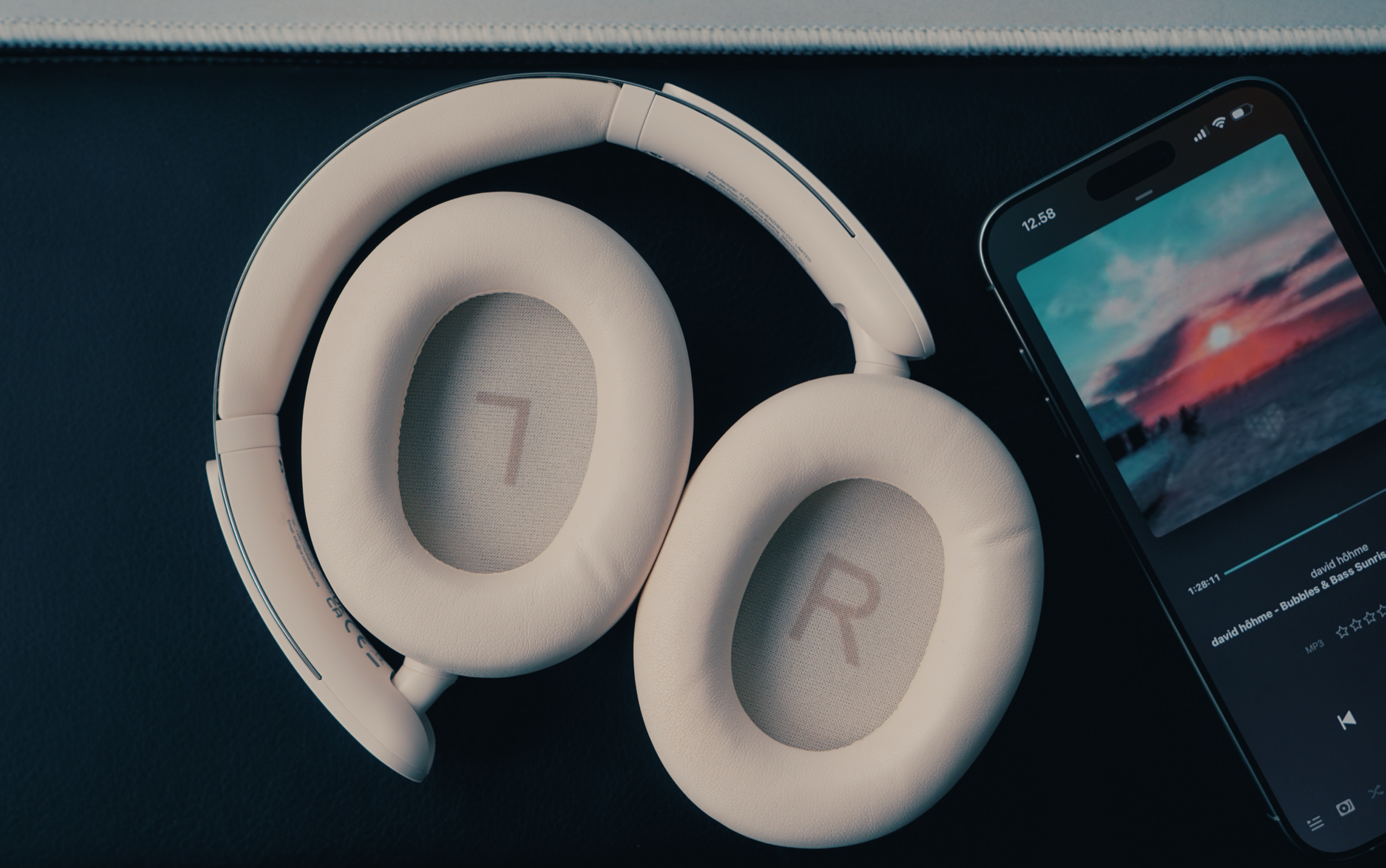 Over-ear, light-colored headphones positioned on a black surface next to a smartphone displaying a sunset image and music track information. The headphones have "L" and "R" markings on their earcups.
