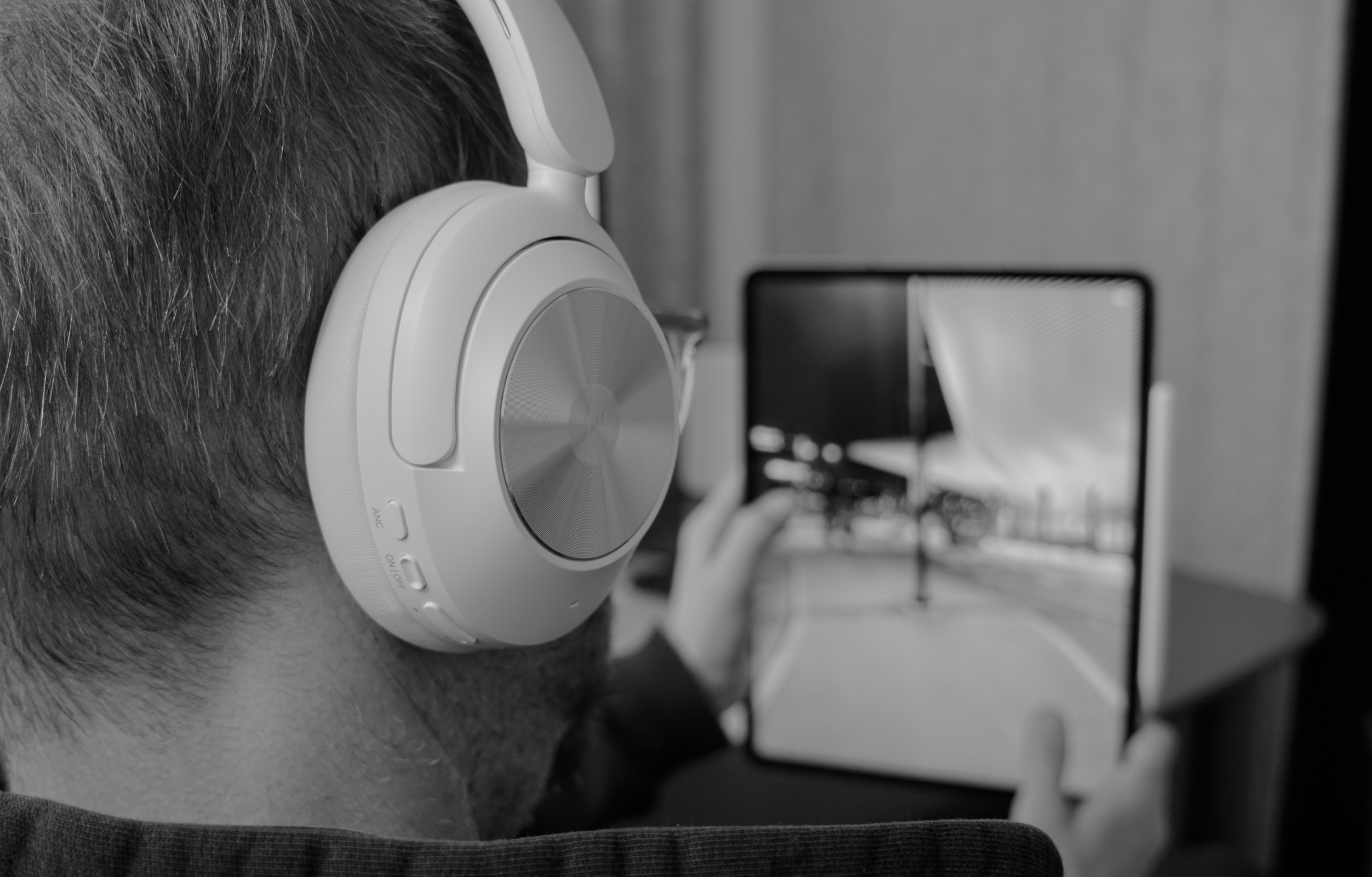 Man wearing white over-ear headphones, seated and viewing a tablet with a cityscape image.
