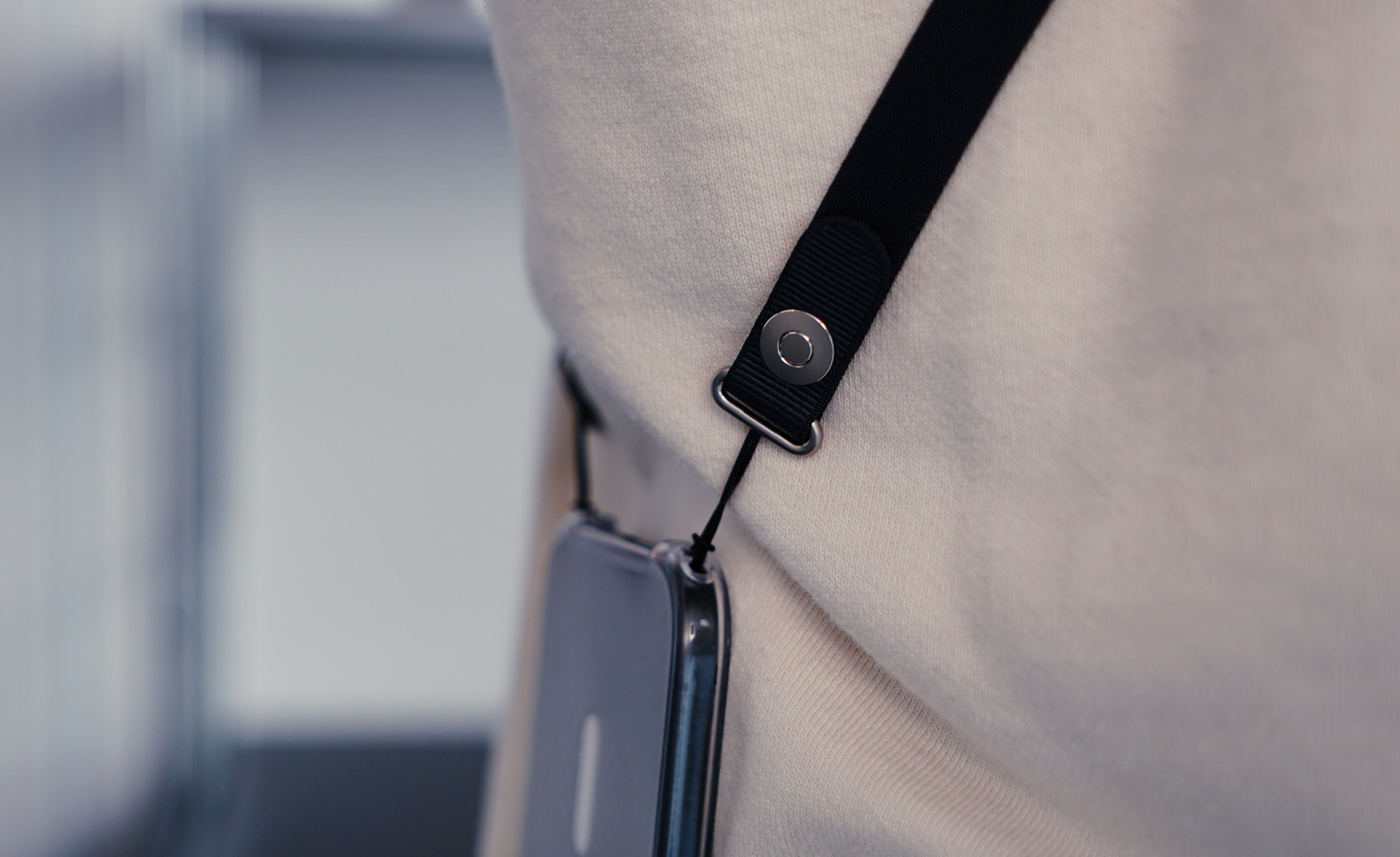Close-up of a cream-colored hoodie with a black lanyard strap attached. The strap is connected to a clear phone case with a black string, the strap has a metal clasp and a silver button. The background is a blurred, light-colored wall.
