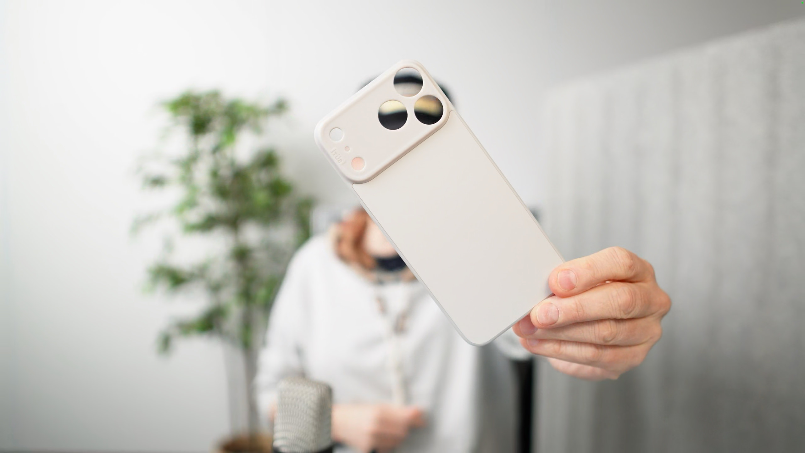 A person holding a white phone case with camera cutouts towards the camera, with blurred background featuring a person, microphone and leafy plant.
