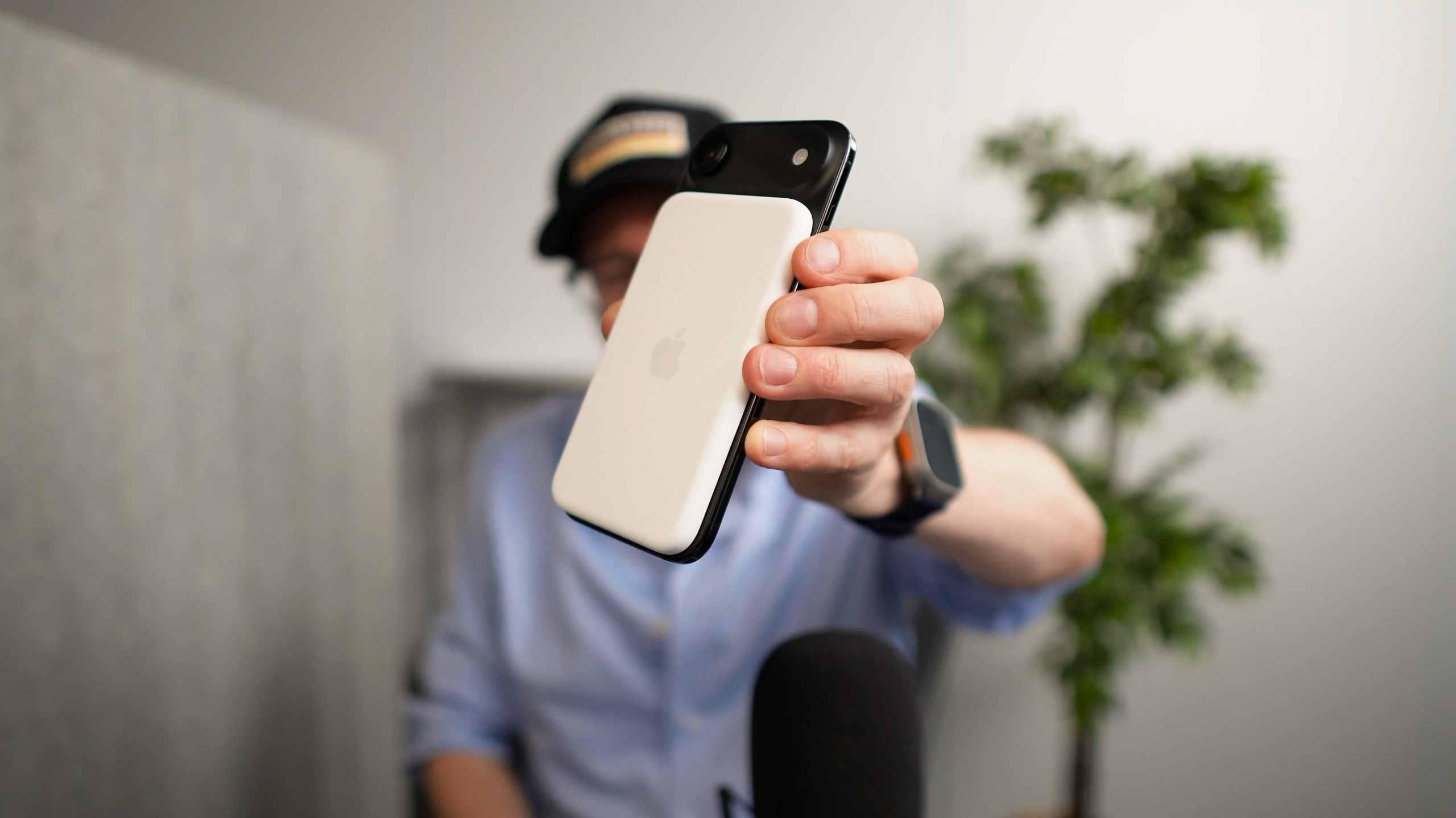 A person, wearing a hat and glasses, holding up a white and black smartphone with the Apple logo visible on its back; background includes a blurred plant and a microphone.
