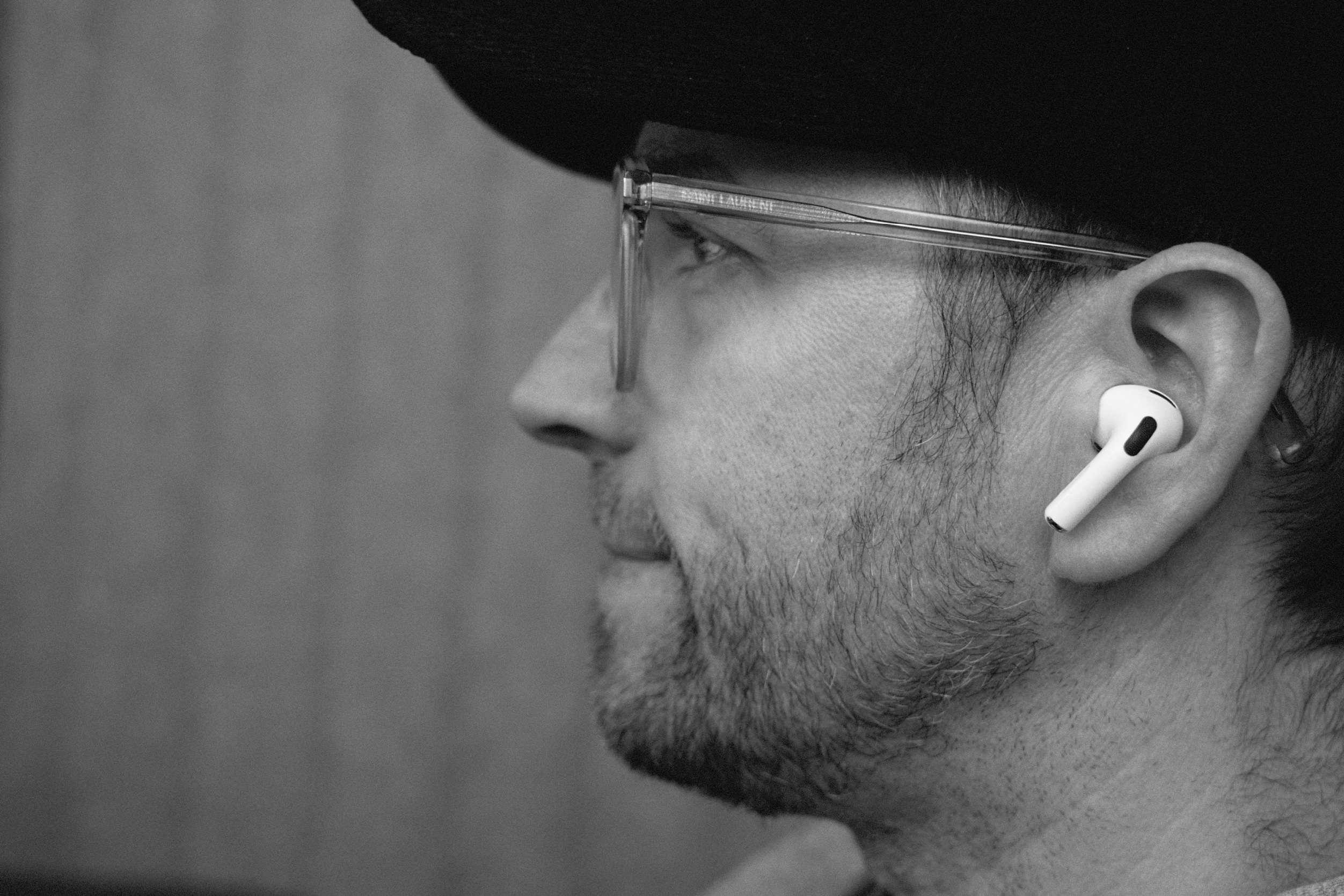 Black and white profile portrait of a man with a beard and glasses wearing a white wireless earbud.
