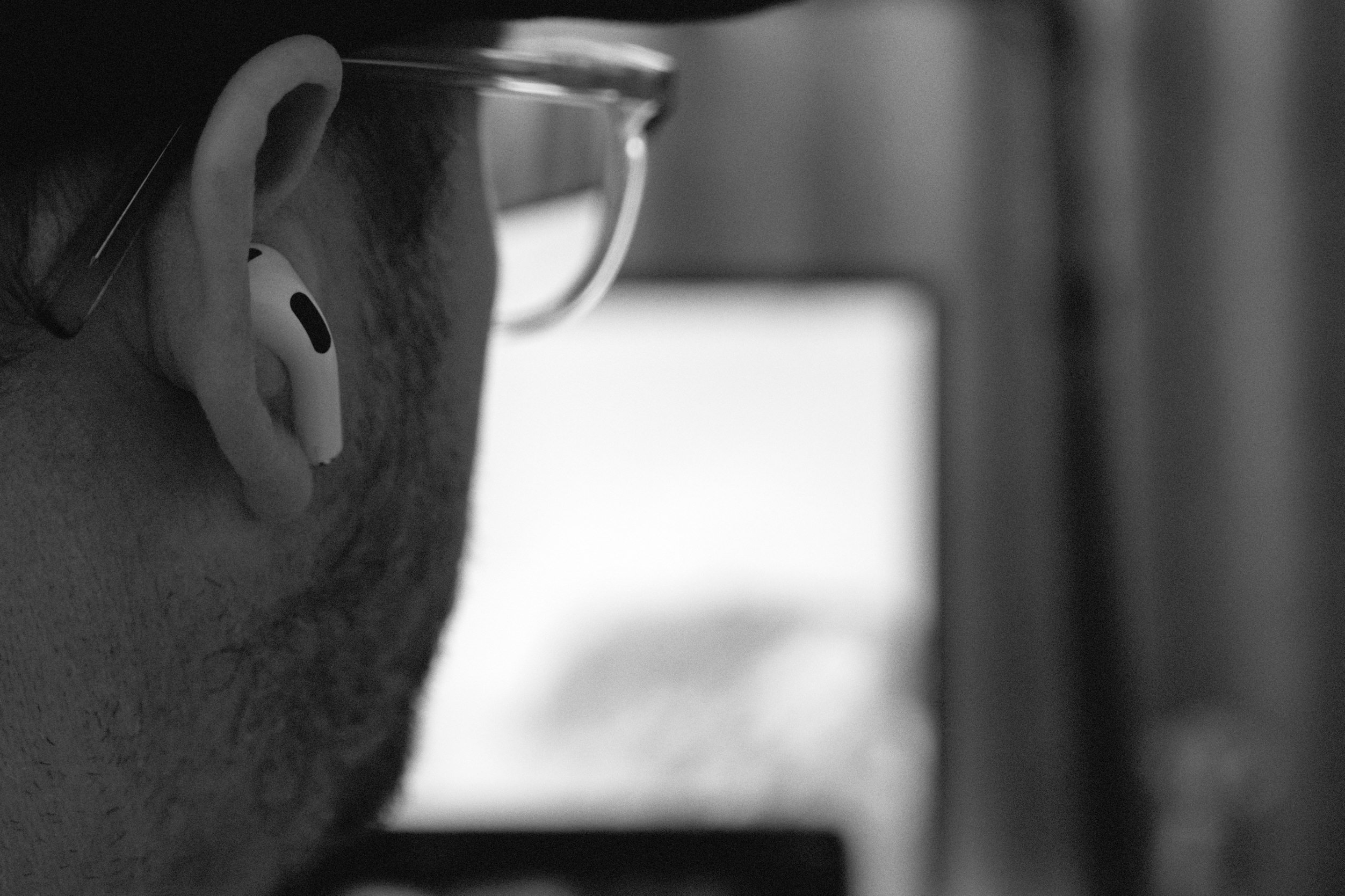 Close-up, black and white shot of a person from behind, wearing glasses and an earbud in their left ear. The focus is on the ear and the white earbud, with a blurred background showing what appears to be a computer screen.
