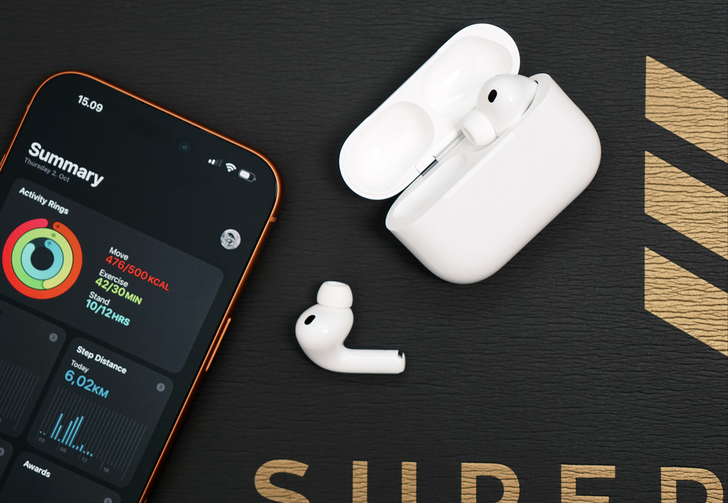 A top-down shot features an open white earbud charging case with an earbud inside, a single white earbud beside the open case, and a smartphone with an orange border displaying health data, all placed on a black textured surface. The phone's screen shows activity rings, step distance, and other exercise metrics. The word "SUPER" is visible in the bottom part of the frame, with the "D" and "E" also visible.
